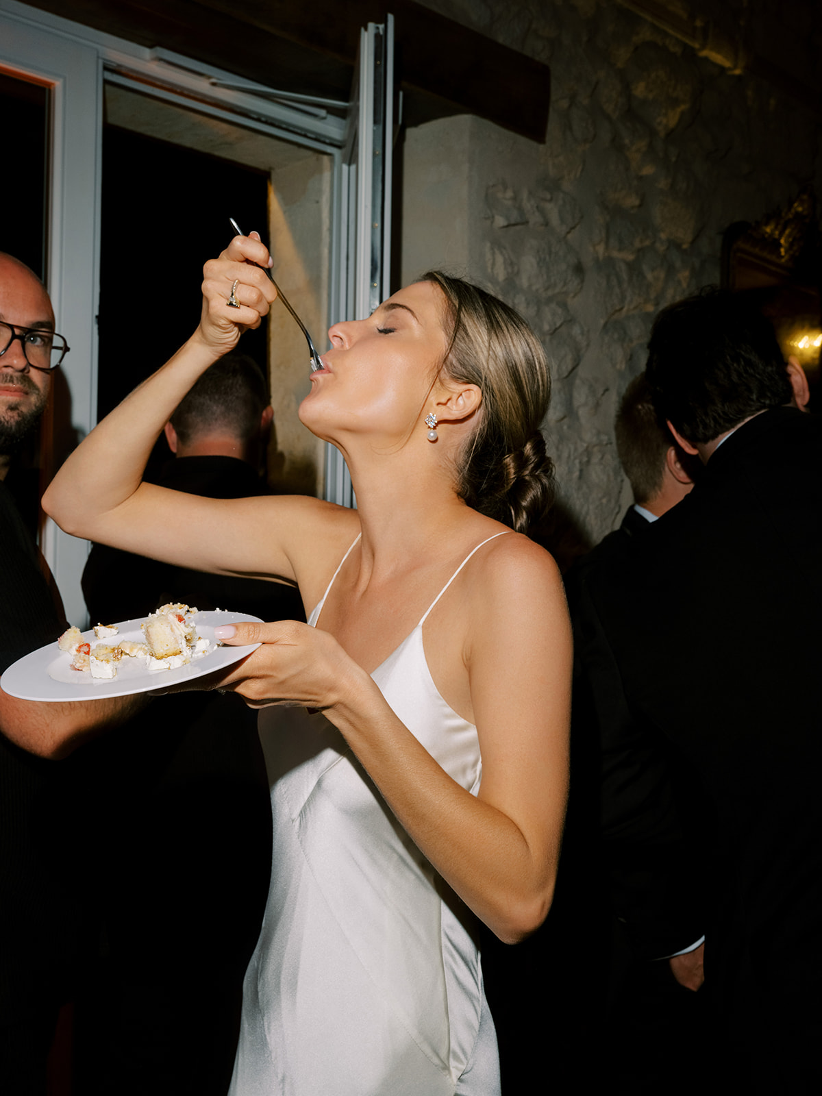 Bride in white slip dress eating from a plate held by a guest during an indoor reception; sage-green walls in background.