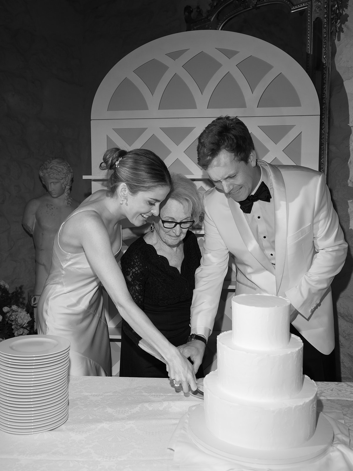 Black-and-white photo of bride, groom, and older guest cutting three-tiered white cake in arched reception room