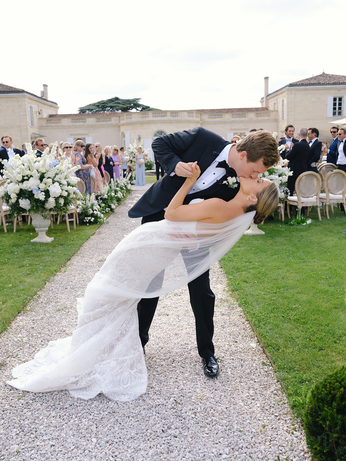 Groom dipping bride on gravel aisle with hydrangea urns and neoclassical chateau backdrop