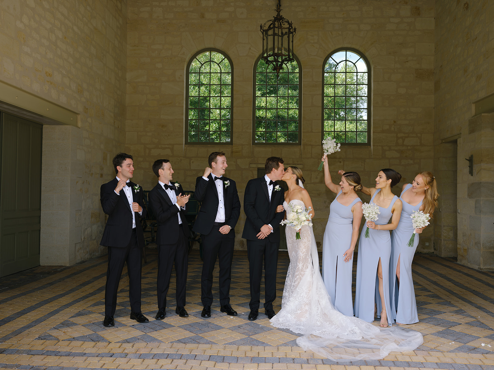Full bridal party portrait in stone hall with groomsmen in black tuxedos and bridesmaids in periwinkle dresses