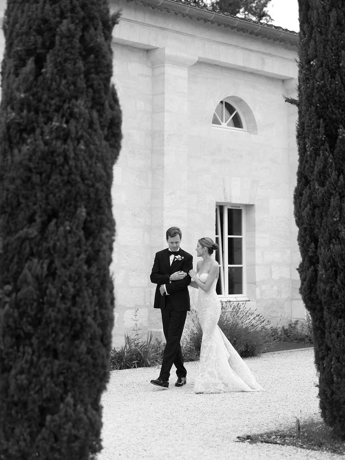 Black and white portrait of couple in a gravel courtyard framed by cypress trees and a white stone chapel