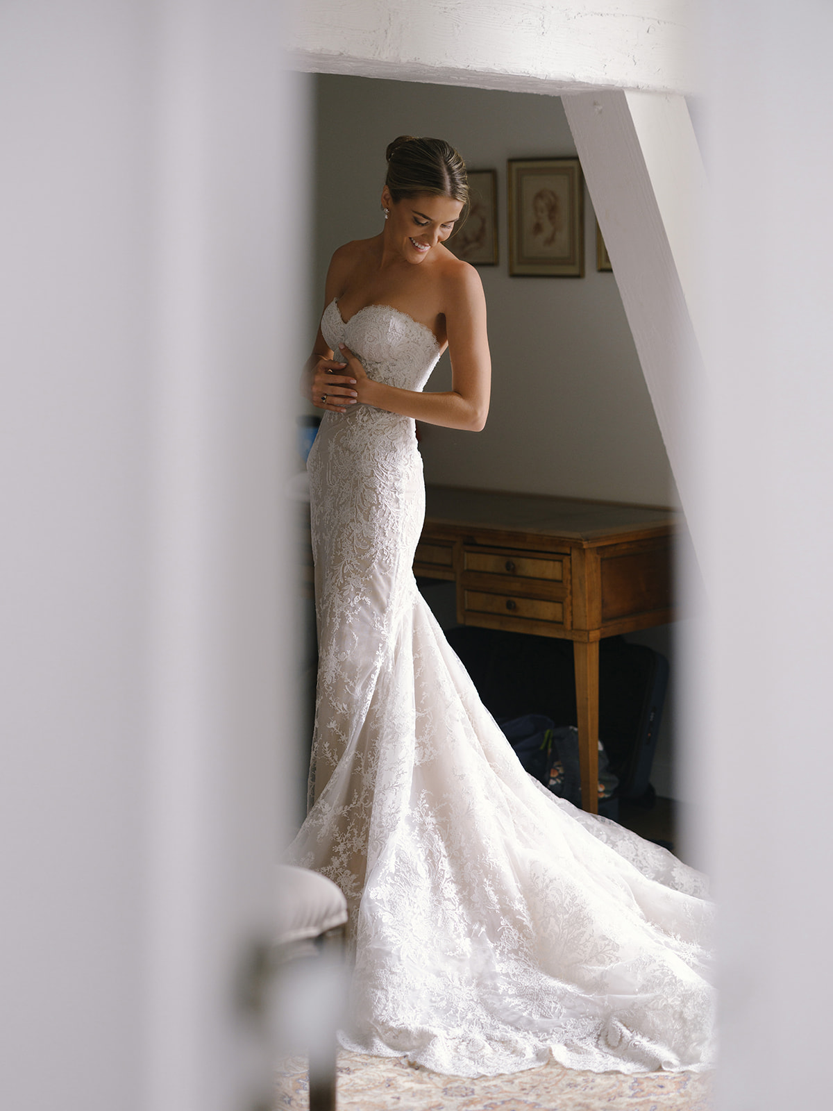 Bride in ivory mermaid gown with beaded lace bodice before large mirror, sleek updo, gazing downward with natural smile