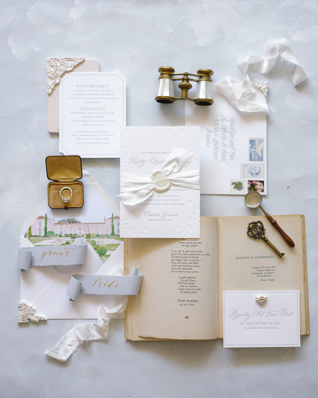 Wedding stationery flat lay with calligraphy invitation, chÃ¢teau illustration, ring in brass box, and vintage opera binoculars