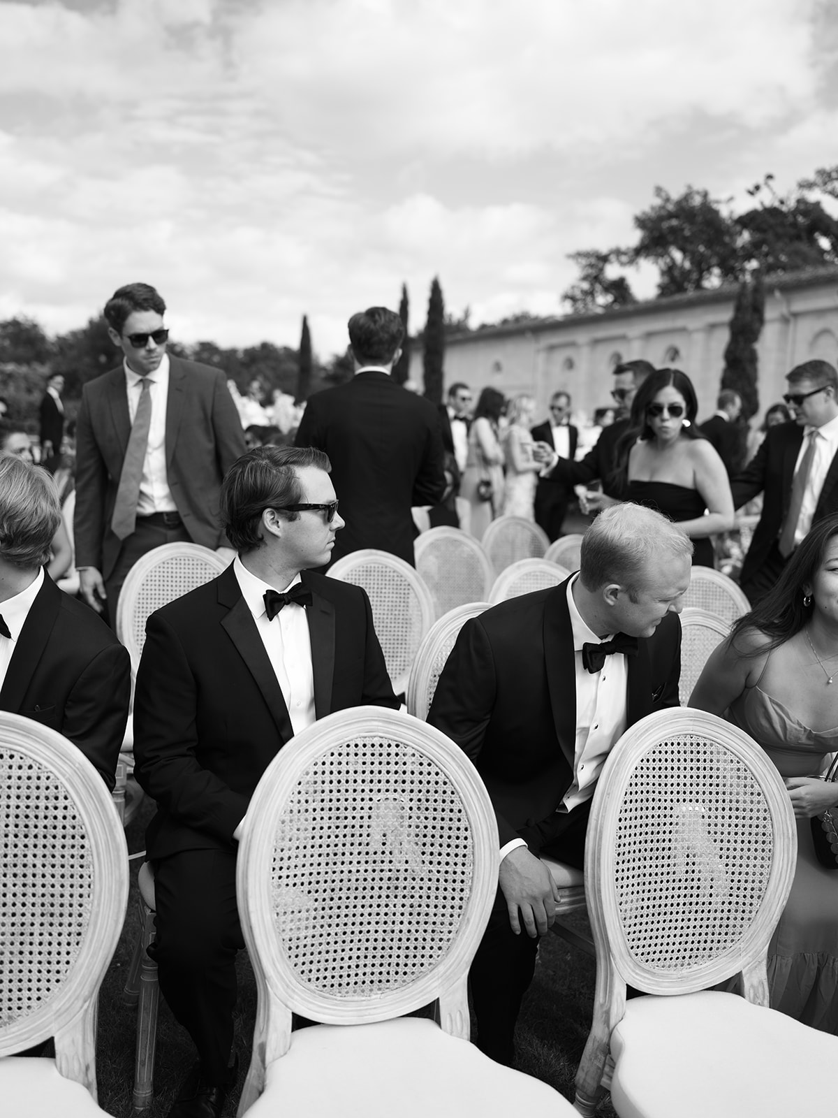 Black-and-white wide shot of guests mingling during cocktail hour in a formal courtyard with cypress trees