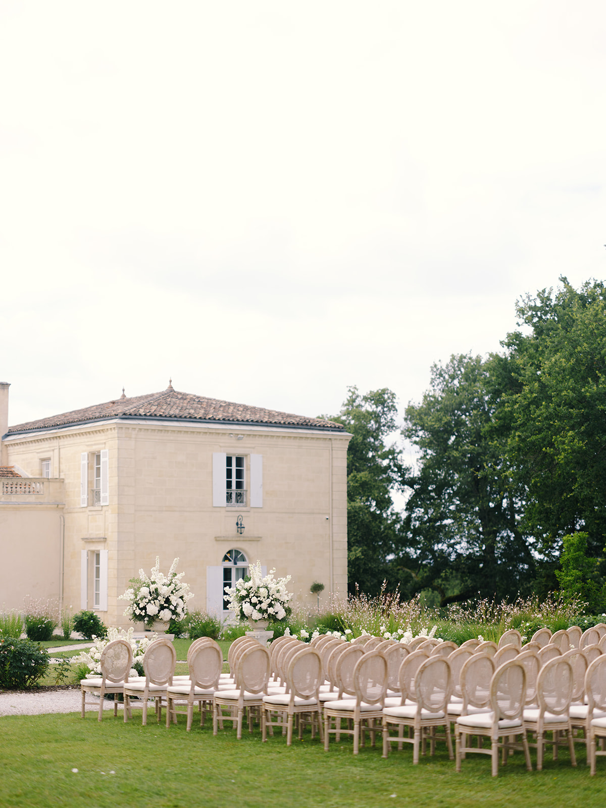 Outdoor ceremony on lawn in front of stone manor with rounded tower, Louis XVI chairs and large white floral arrangements