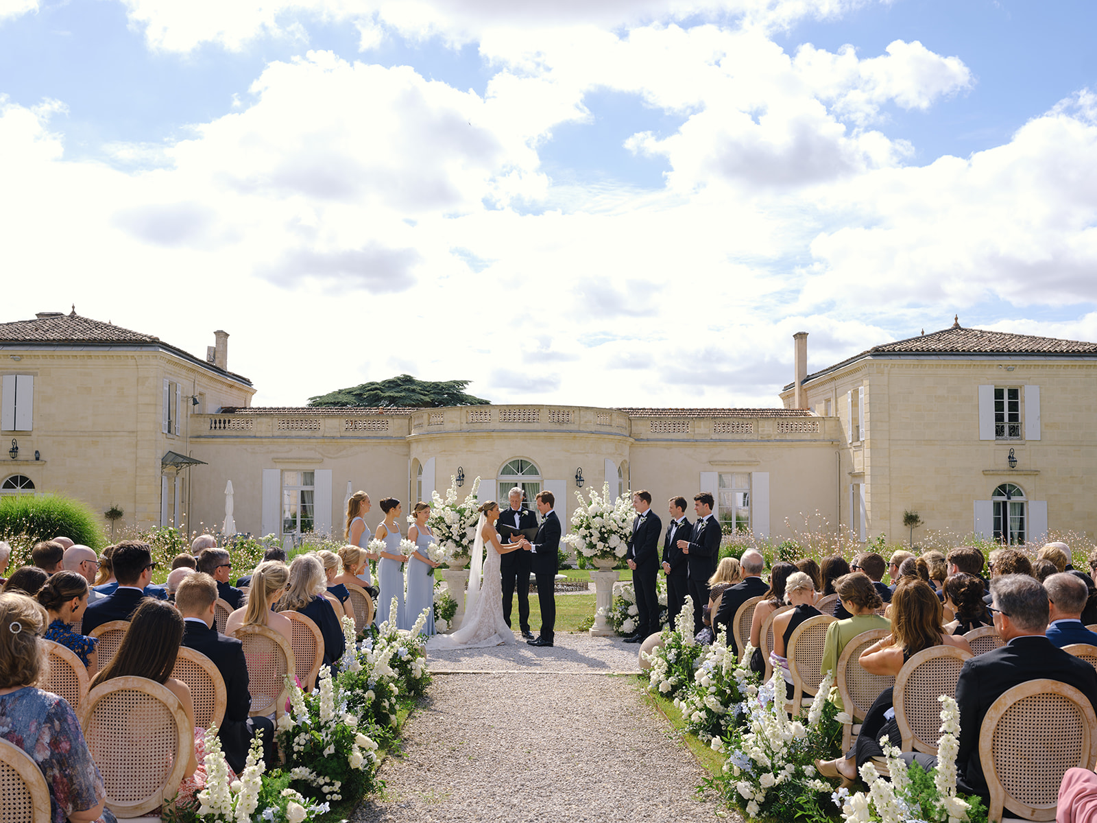 Wide elevated shot of outdoor ceremony in chÃ¢teau courtyard with 80-100 guests and white floral aisle arrangements