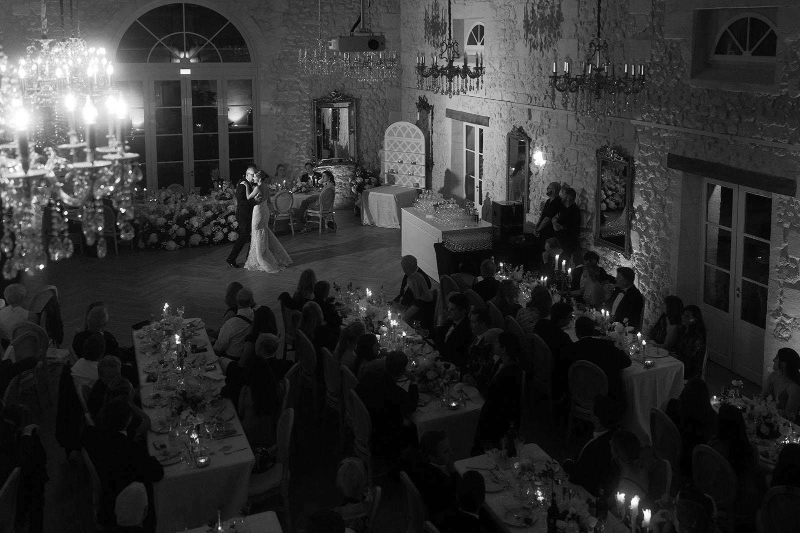 Black and white wide shot of couple dancing in stone ballroom with crystal chandeliers and guests