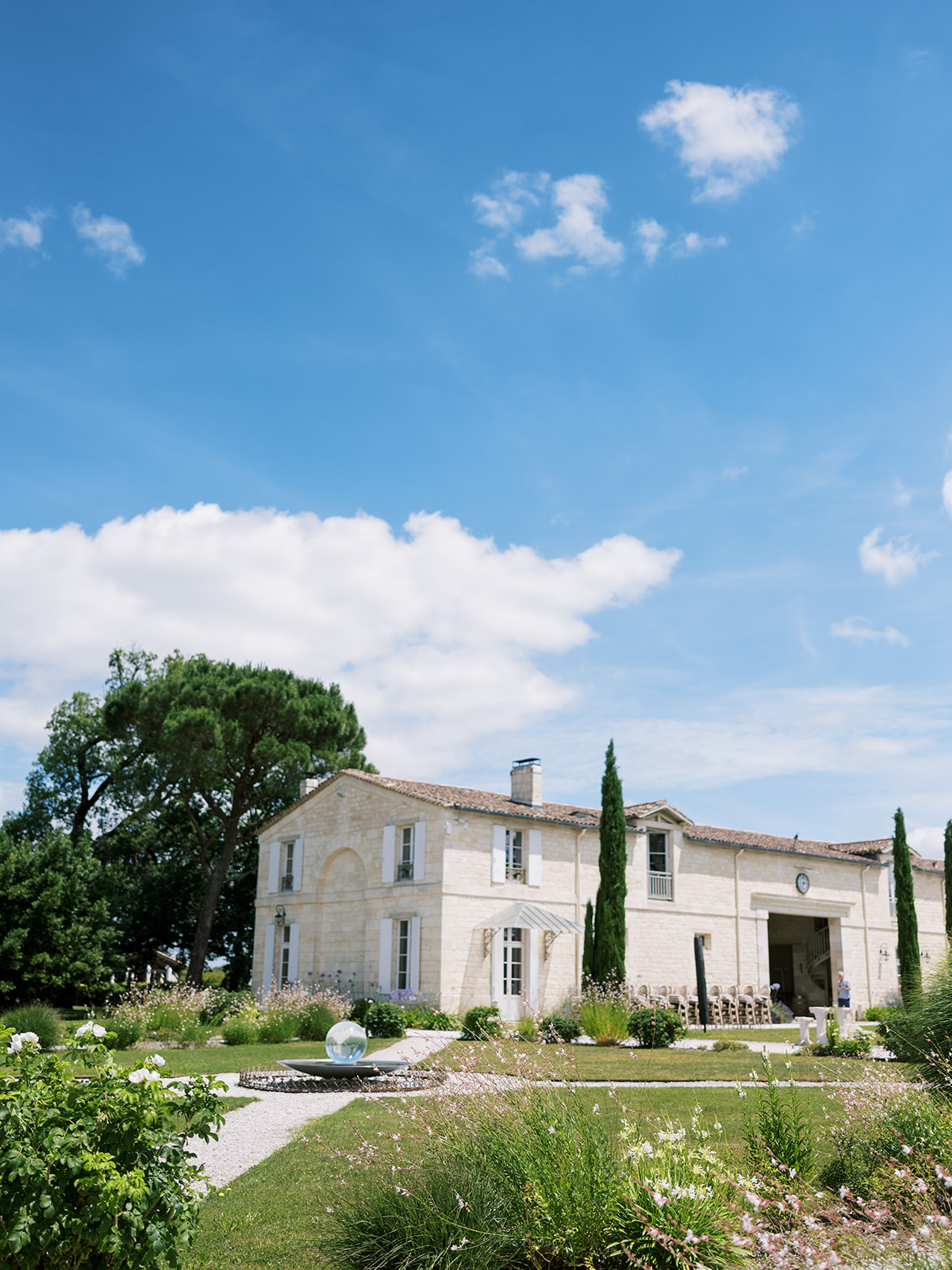 Cream stone chateau under blue sky with cypress trees, garden water feature and guests on formal grounds