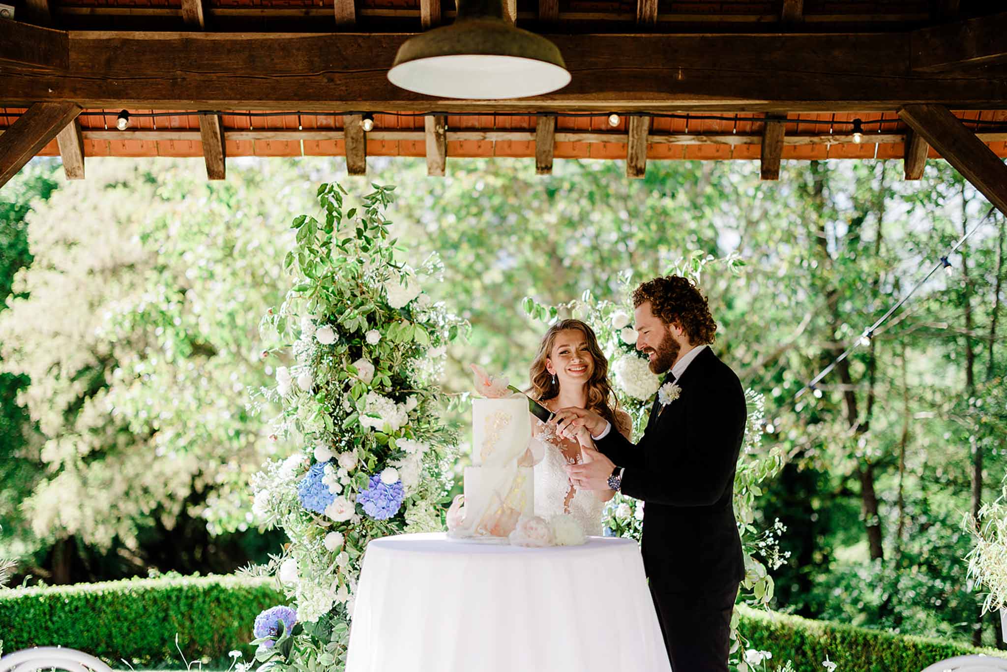 Bride and groom cutting white wedding cake under pergola flanked by white roses, hydrangeas, and eucalyptus floral installations