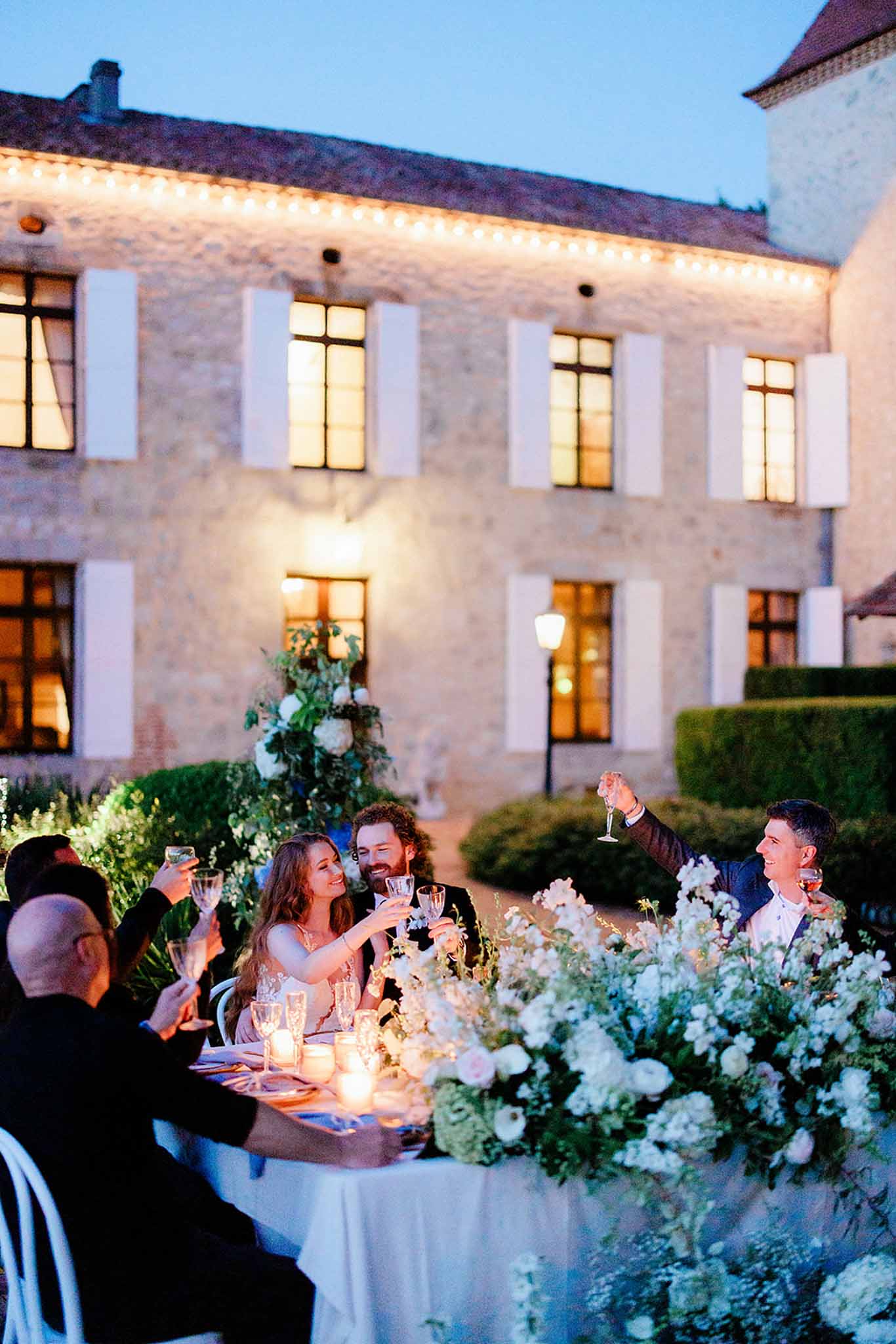 Evening reception dinner in stone courtyard with uplighting, string lights, and white florals