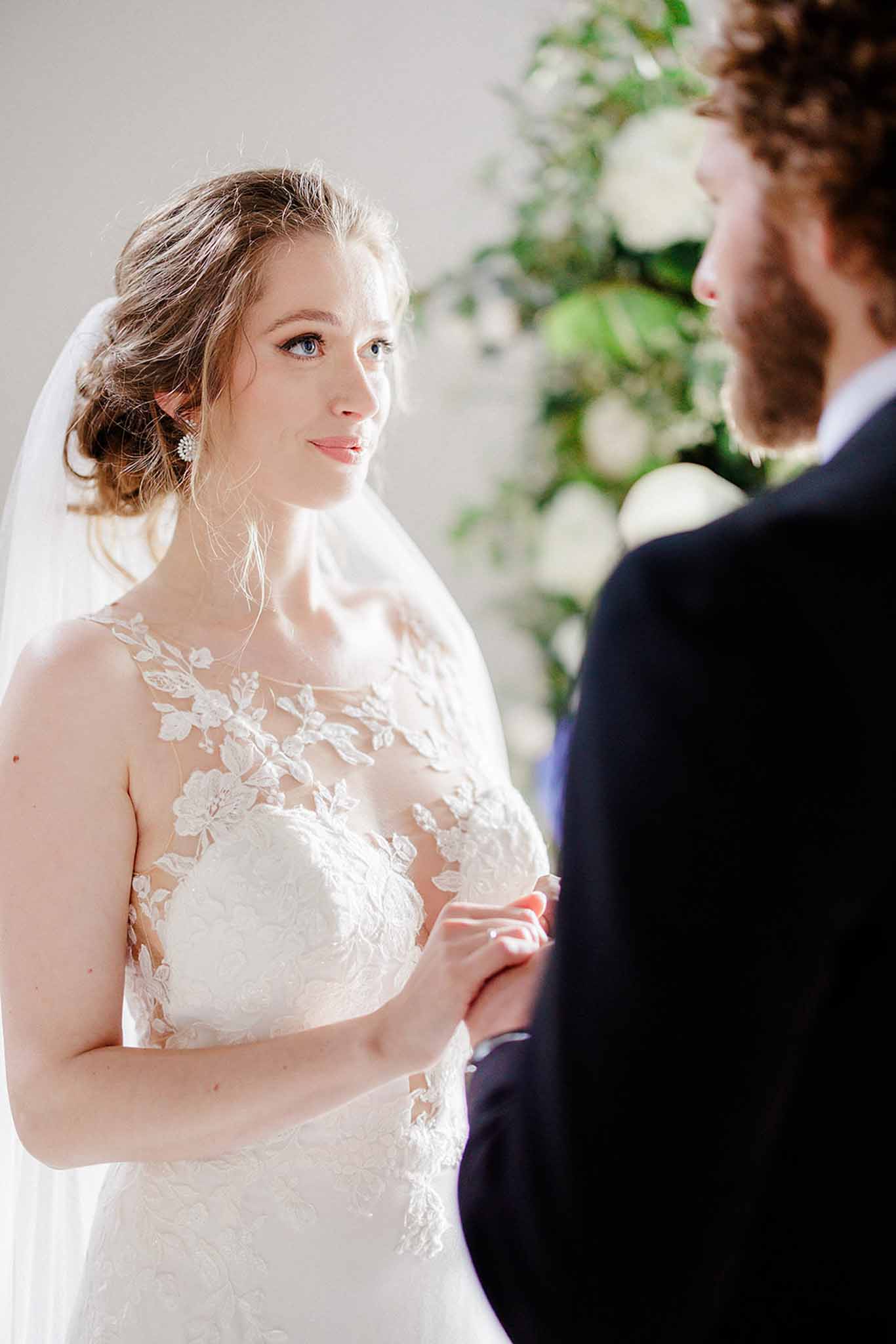 Bride in ivory lace gown with 3D floral appliquÃ©s and groom in navy suit during ceremony vows before white floral backdrop
