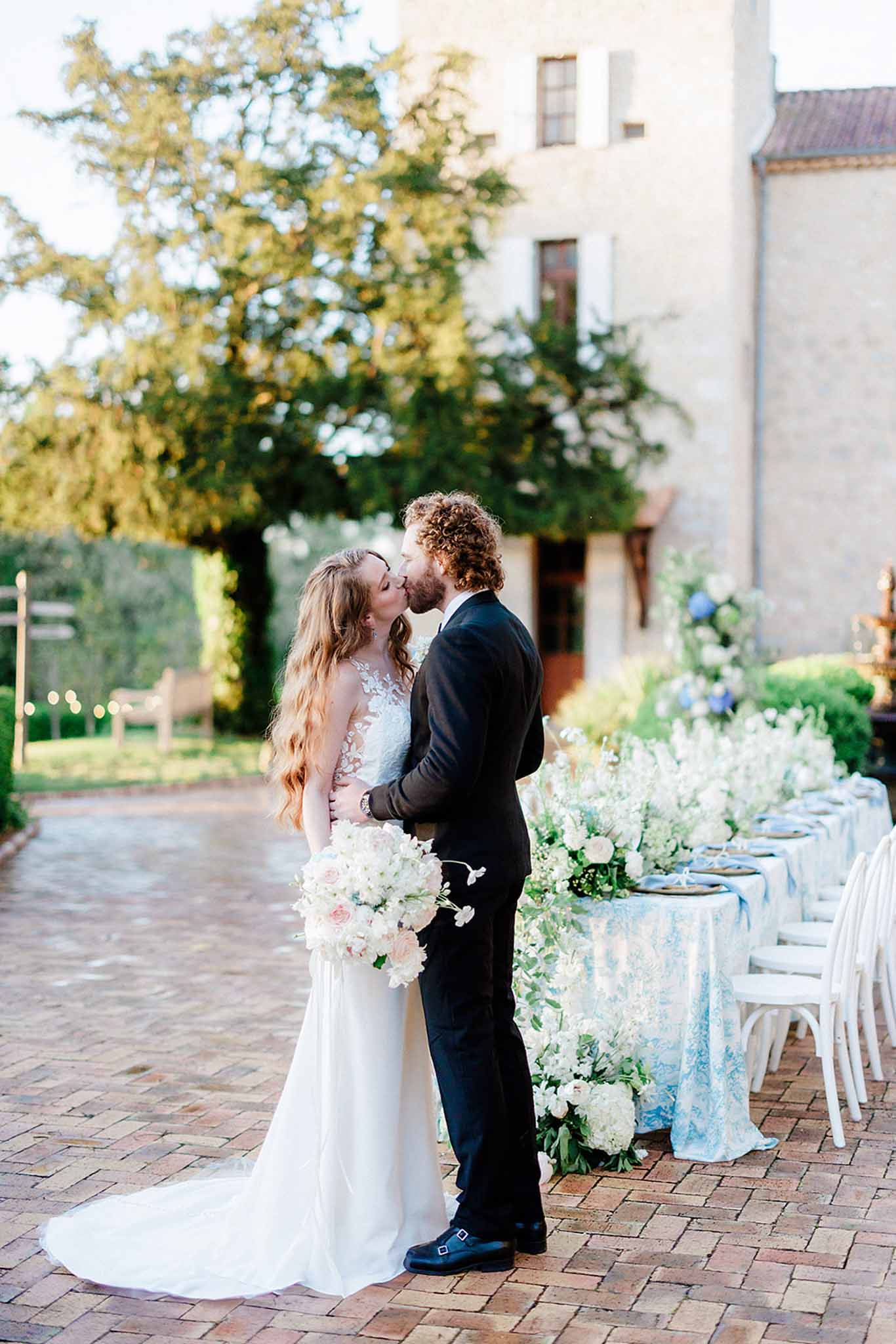 Bride and groom kissing in brick courtyard beside stone building, long dining table with blue linen visible to the right
