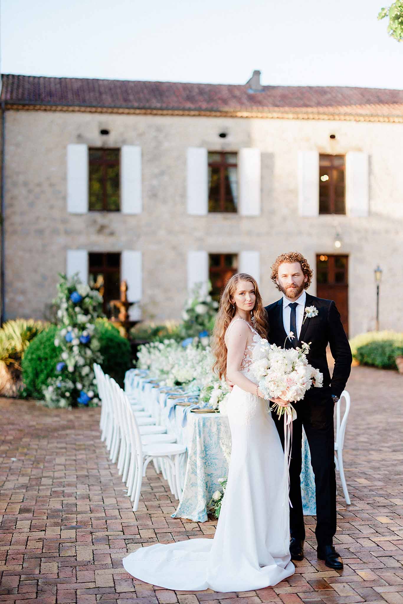 Bride and groom pose in courtyard in front of dressed reception table with blue runners and hydrangea arrangements