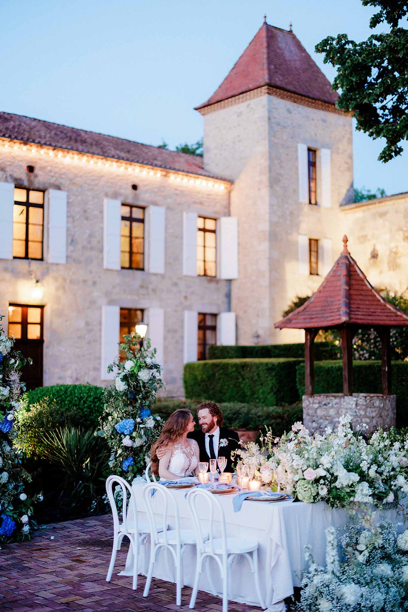 Intimate reception table for two at dusk in chÃ¢teau courtyard with blue delphiniums and lit architectural backdrop