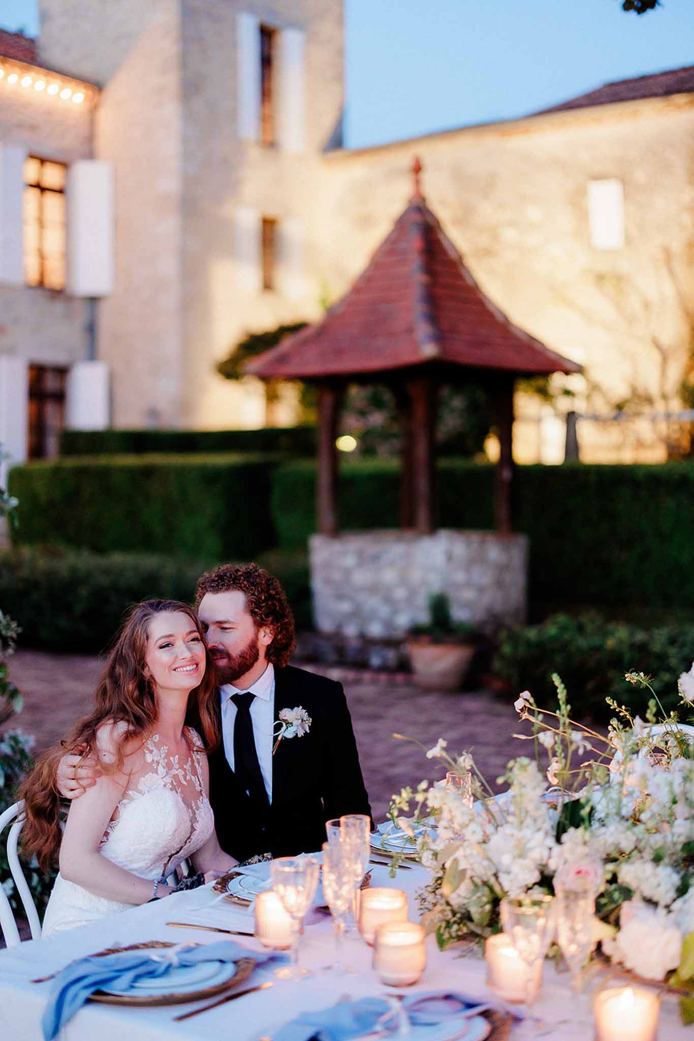 Bride and groom seated at candlelit outdoor reception table in a stone courtyard during twilight