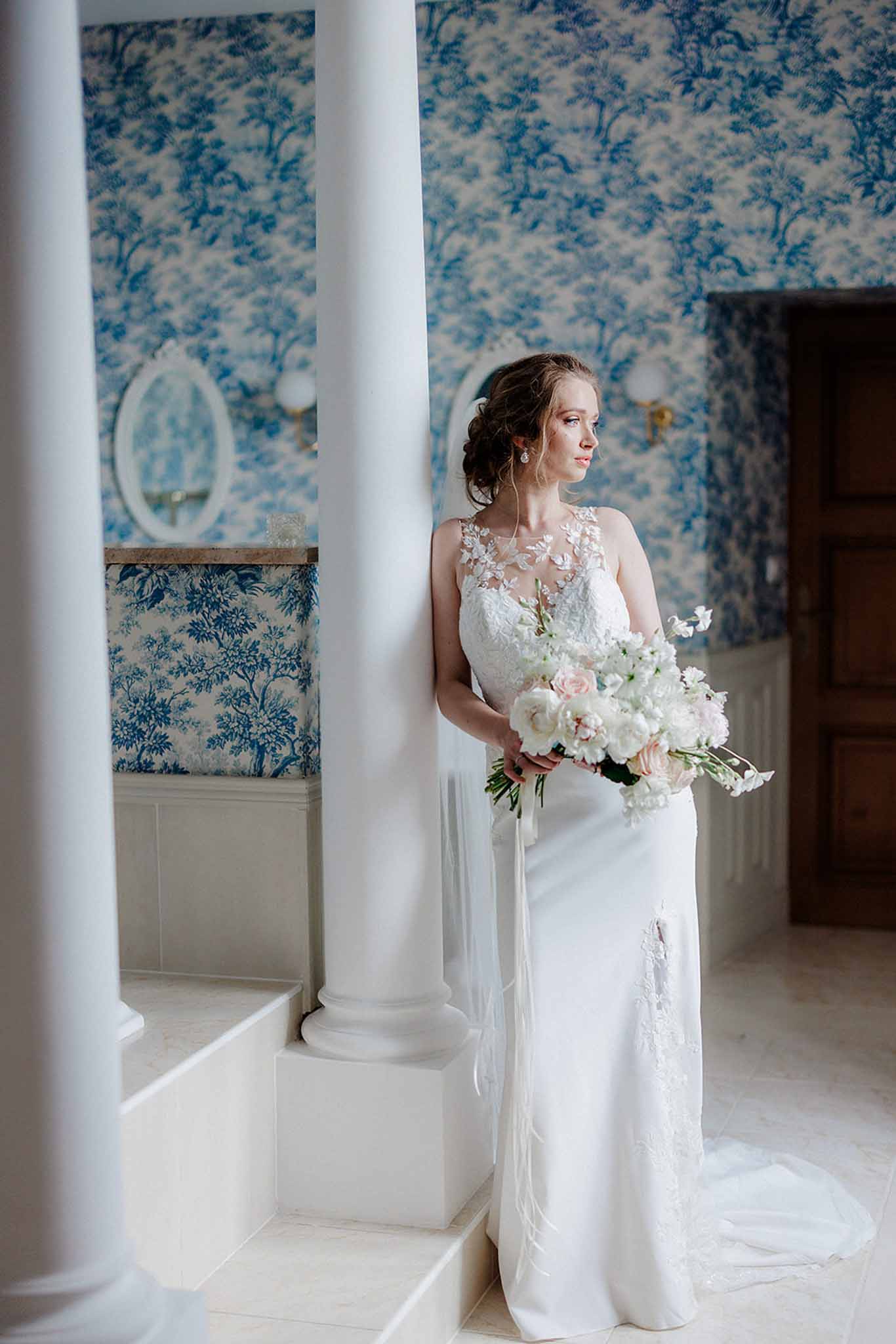 Bride in ivory lace gown holding white and blush rose bouquet between classical white columns with toile wallpaper