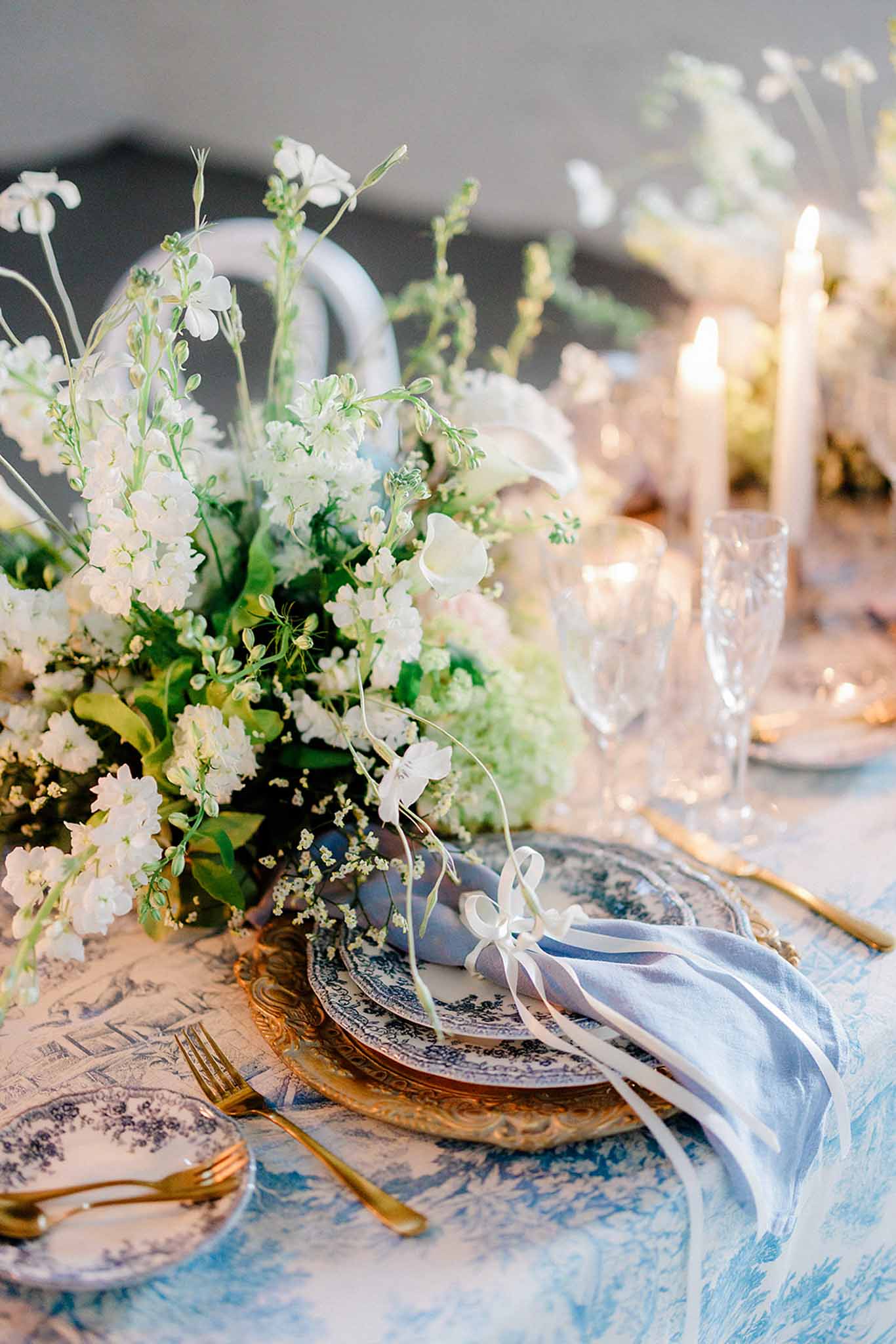 Reception table with blue and white linen, gold flatware, blue-and-white porcelain plates, and white floral centerpieces