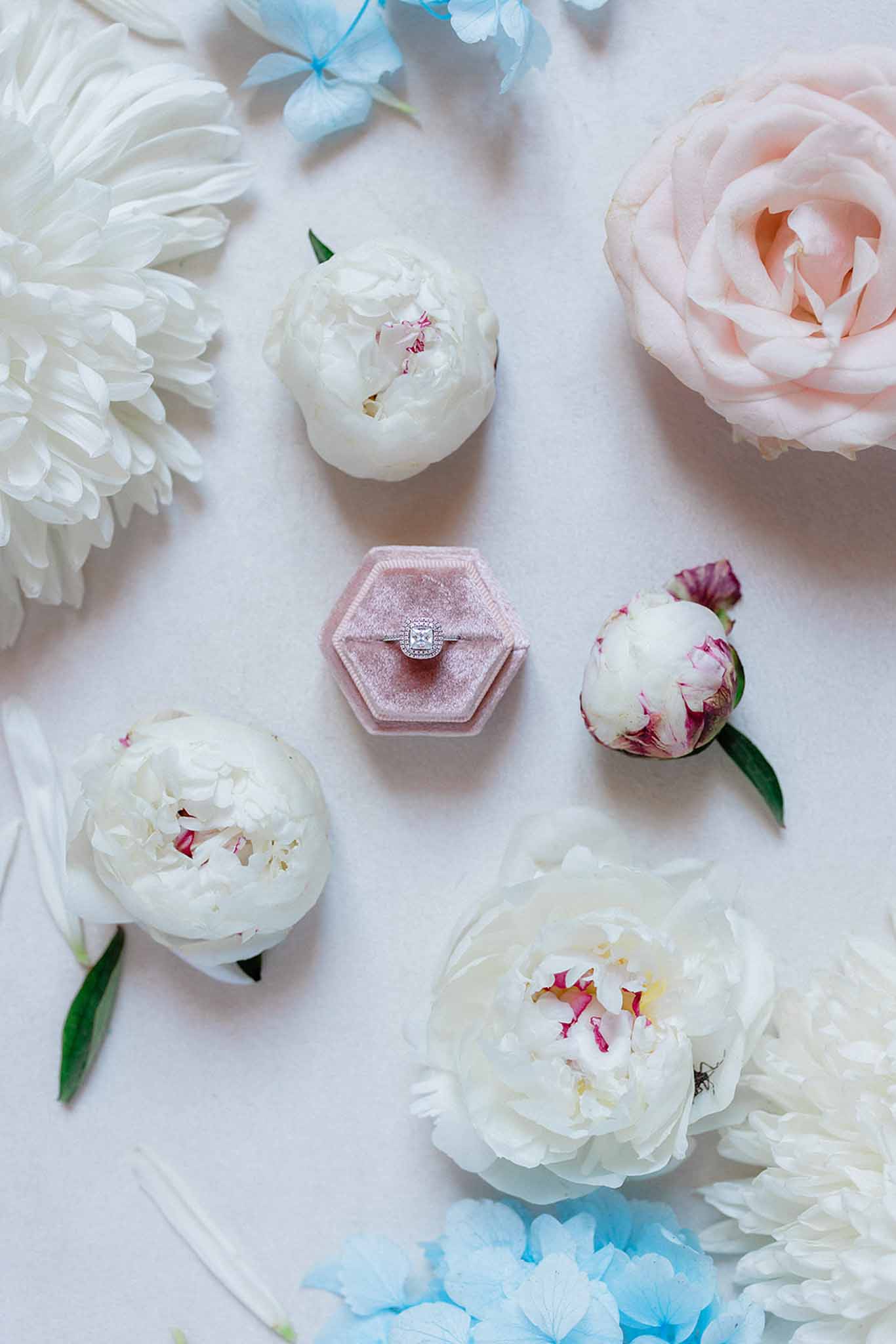 Flat lay of rectangular-cut diamond ring in dusty rose velvet box surrounded by ivory and blush peonies and blue hydrangeas
