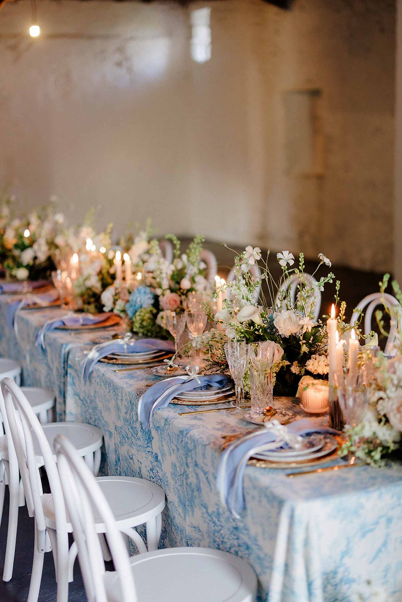 Low-angle view down banquet table with blue and white linen, mixed white florals, candles, and bentwood chairs