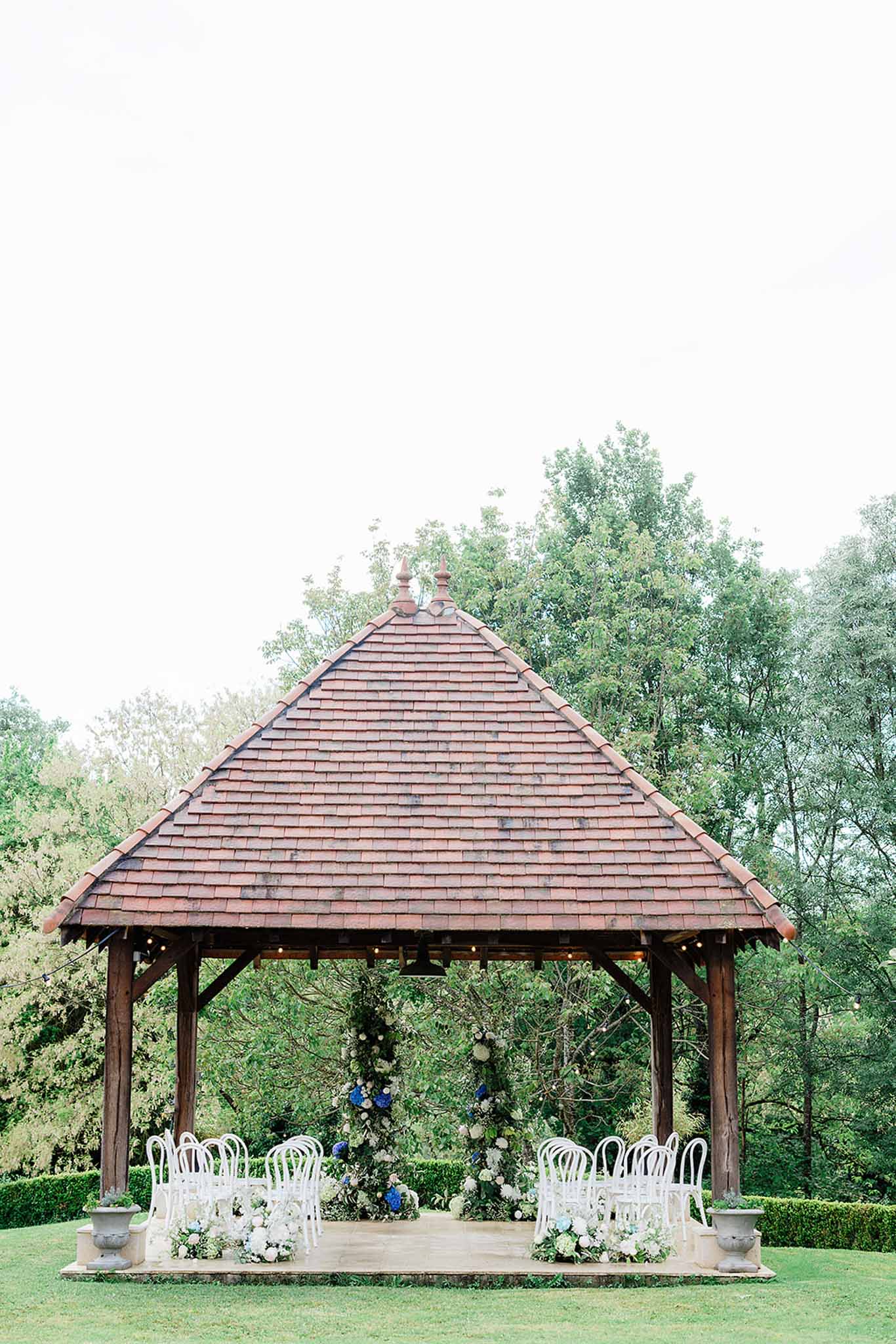 Wooden gazebo with terracotta roof decorated with white and blue florals, white bentwood chairs set for garden ceremony