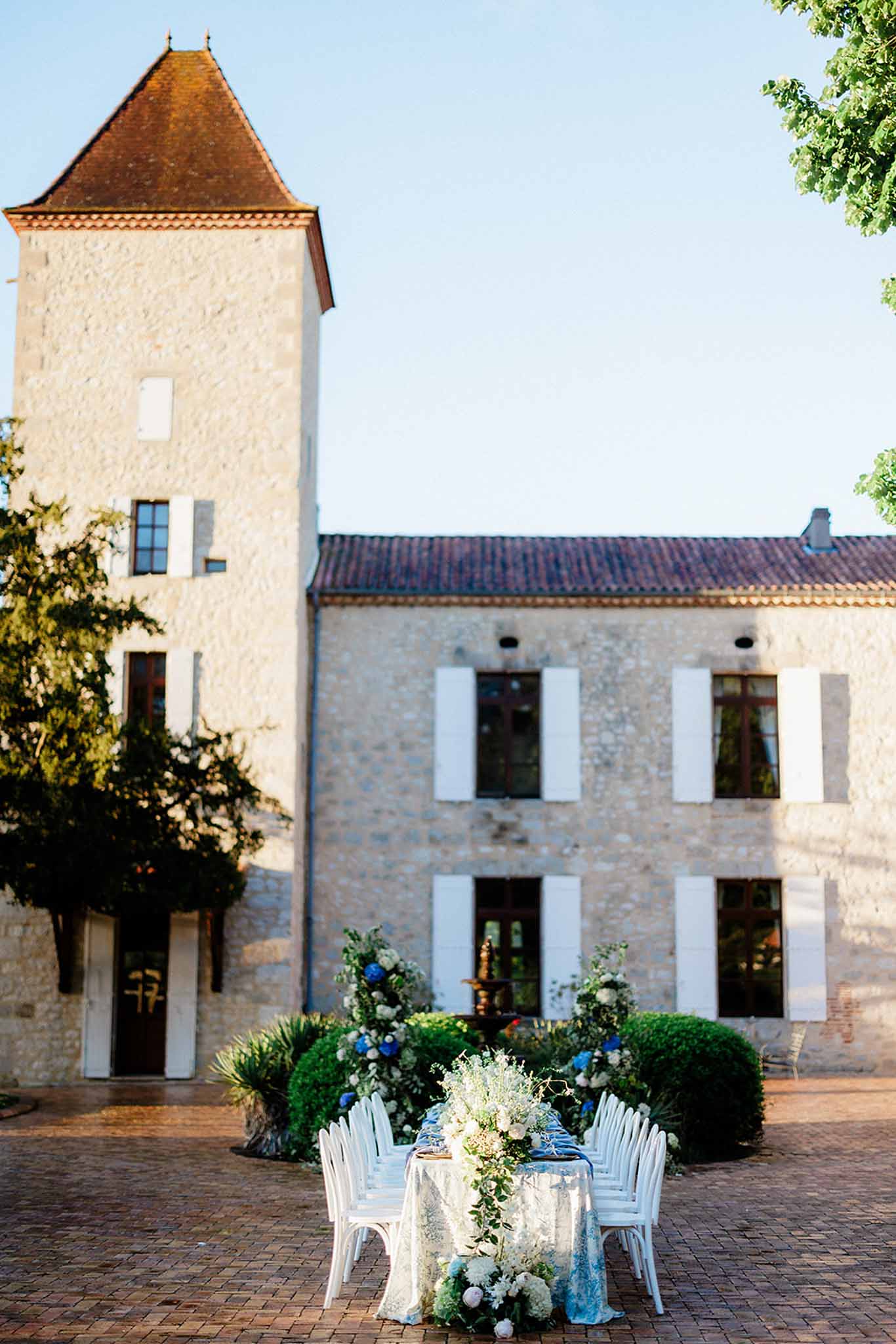 Sweetheart table in courtyard of stone estate with chÃ¢teau tower, white roses and blue hydrangeas in pots