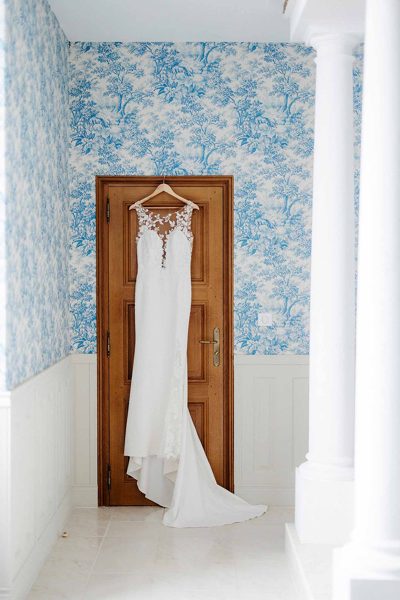 Ivory wedding gown with lace bodice hanging on wooden hanger against blue toile wallpaper