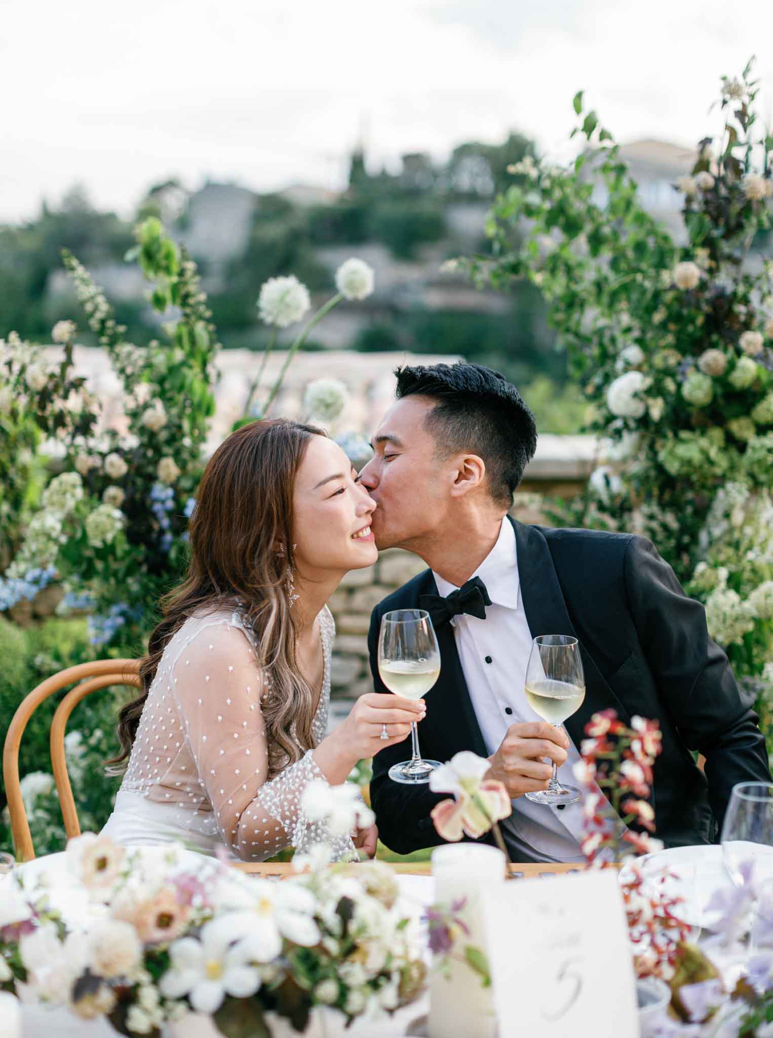 Bride and groom kissing at outdoor reception table with white dahlias and cream roses at hillside venue