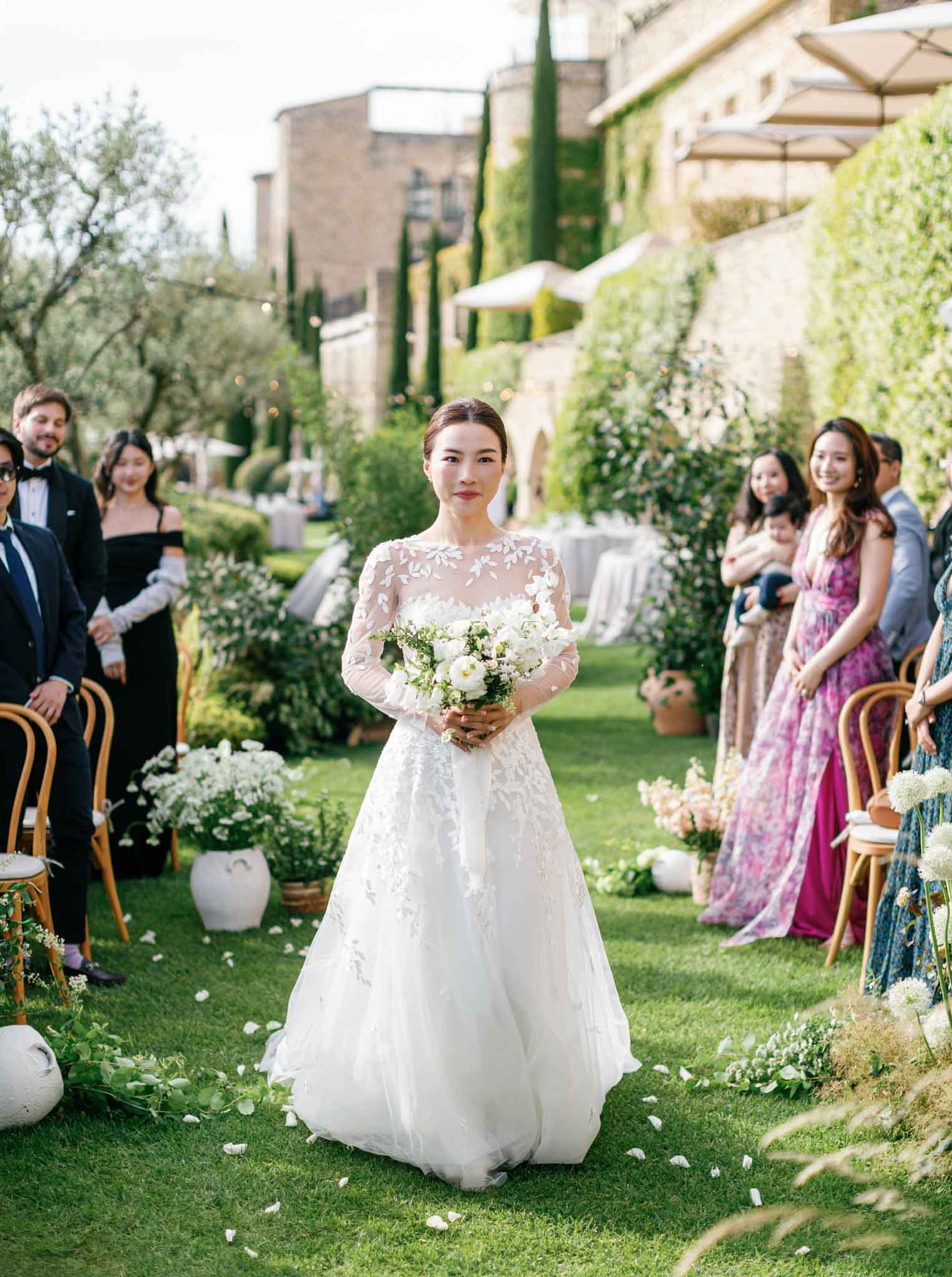 Bride in ivory lace long-sleeve dress walking down outdoor garden aisle lined with seated guests and cream umbrellas