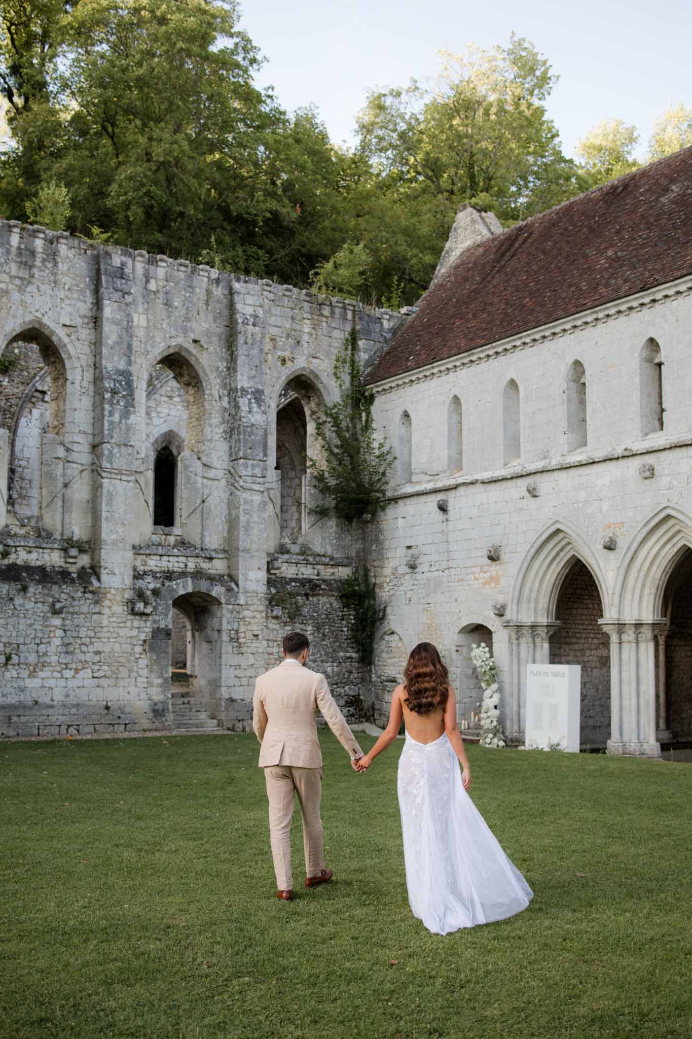 Bride and groom walking across lawn courtyard within ruins of a medieval stone abbey with Gothic arch windows