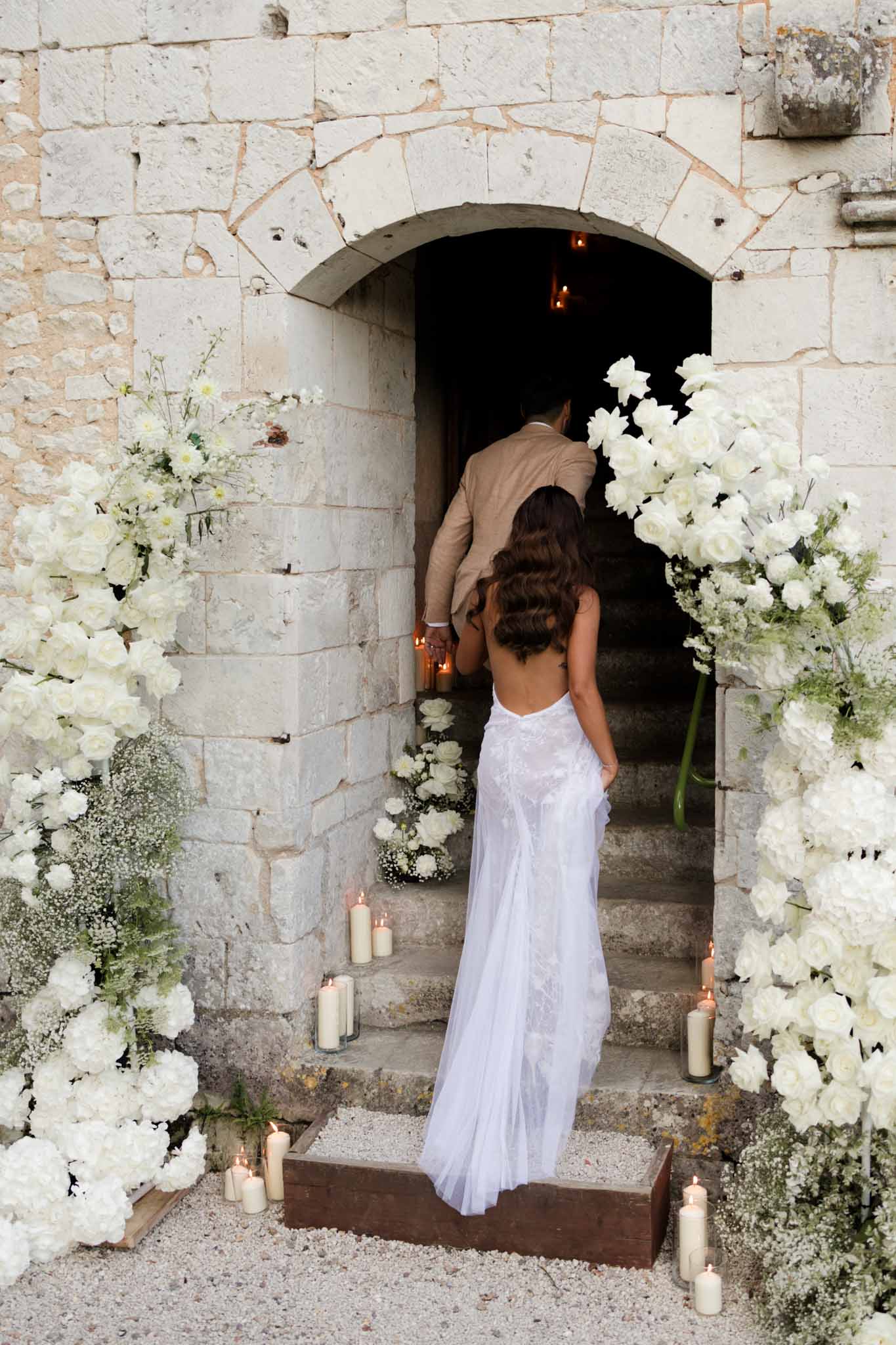 Bride and groom entering stone archway lit by candles with white peony and rose floral arrangements