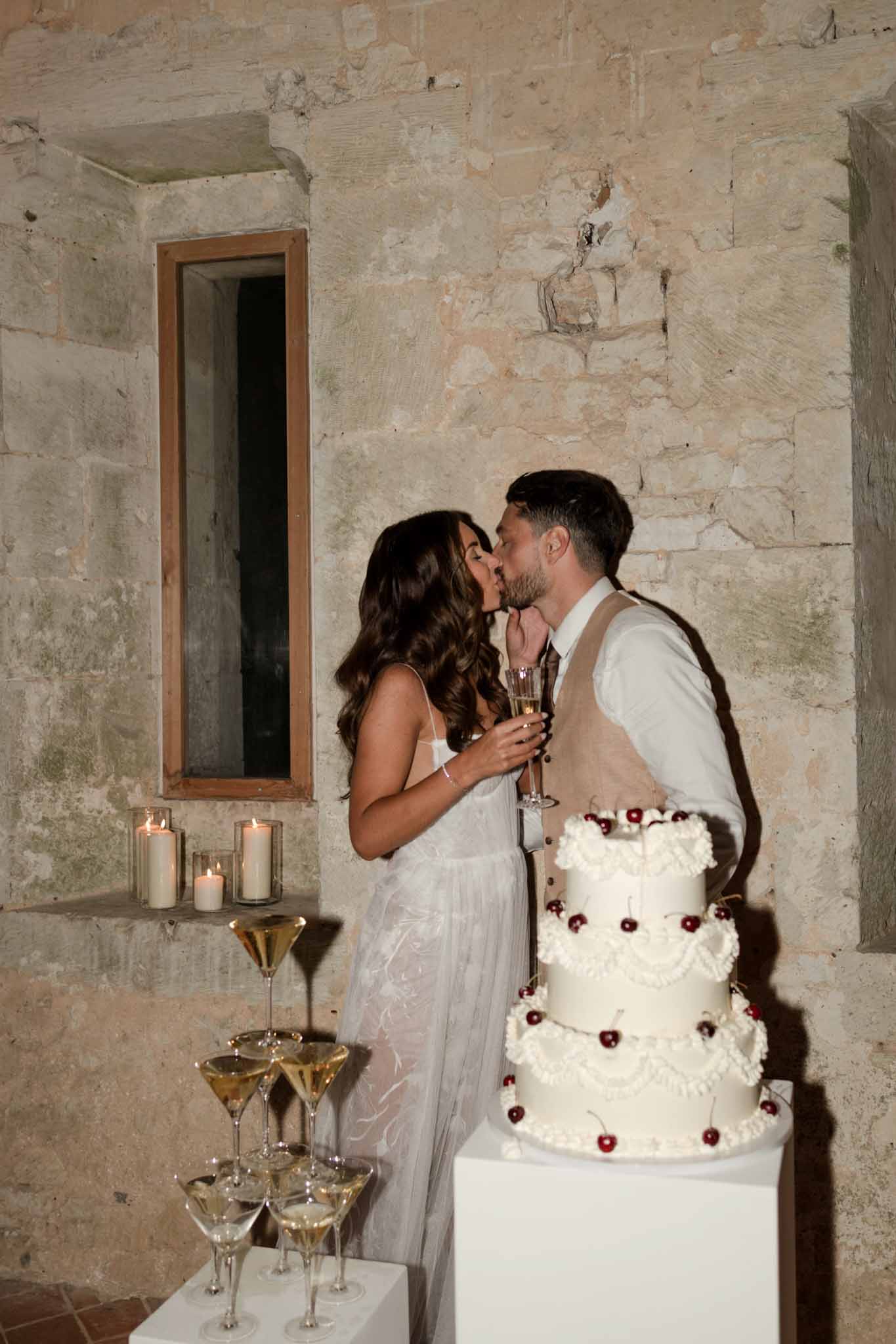 Bride and groom kissing beside three-tier cake with burgundy berries and champagne coupe display