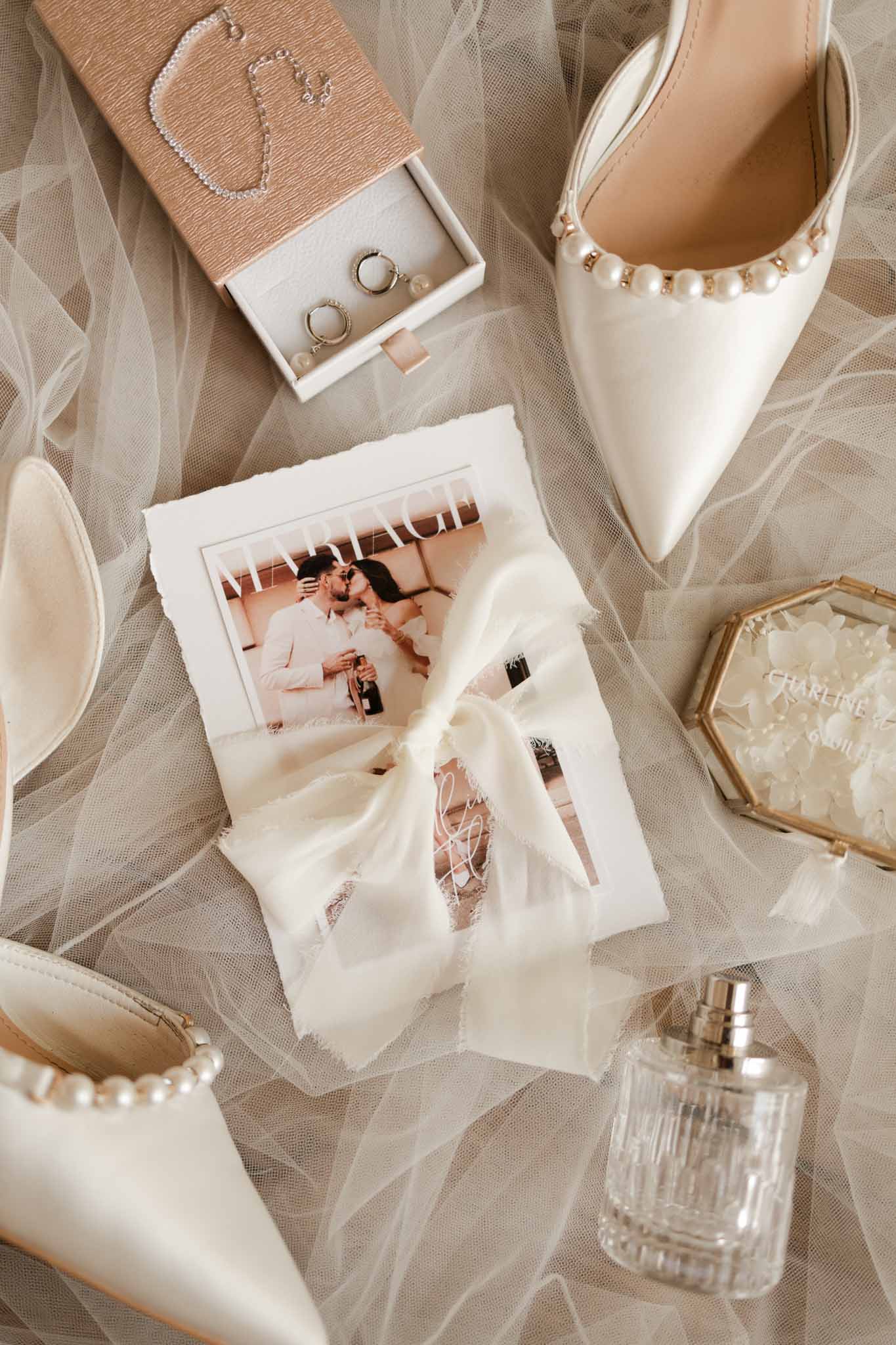 Flat lay of bridal accessories including pearl-beaded heels, silver rings, pearl necklace, and perfume on tulle
