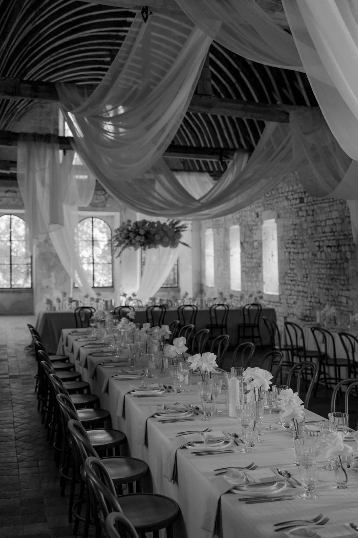 Black-and-white long reception tables in industrial venue with exposed beams, draped fabric, and arched windows