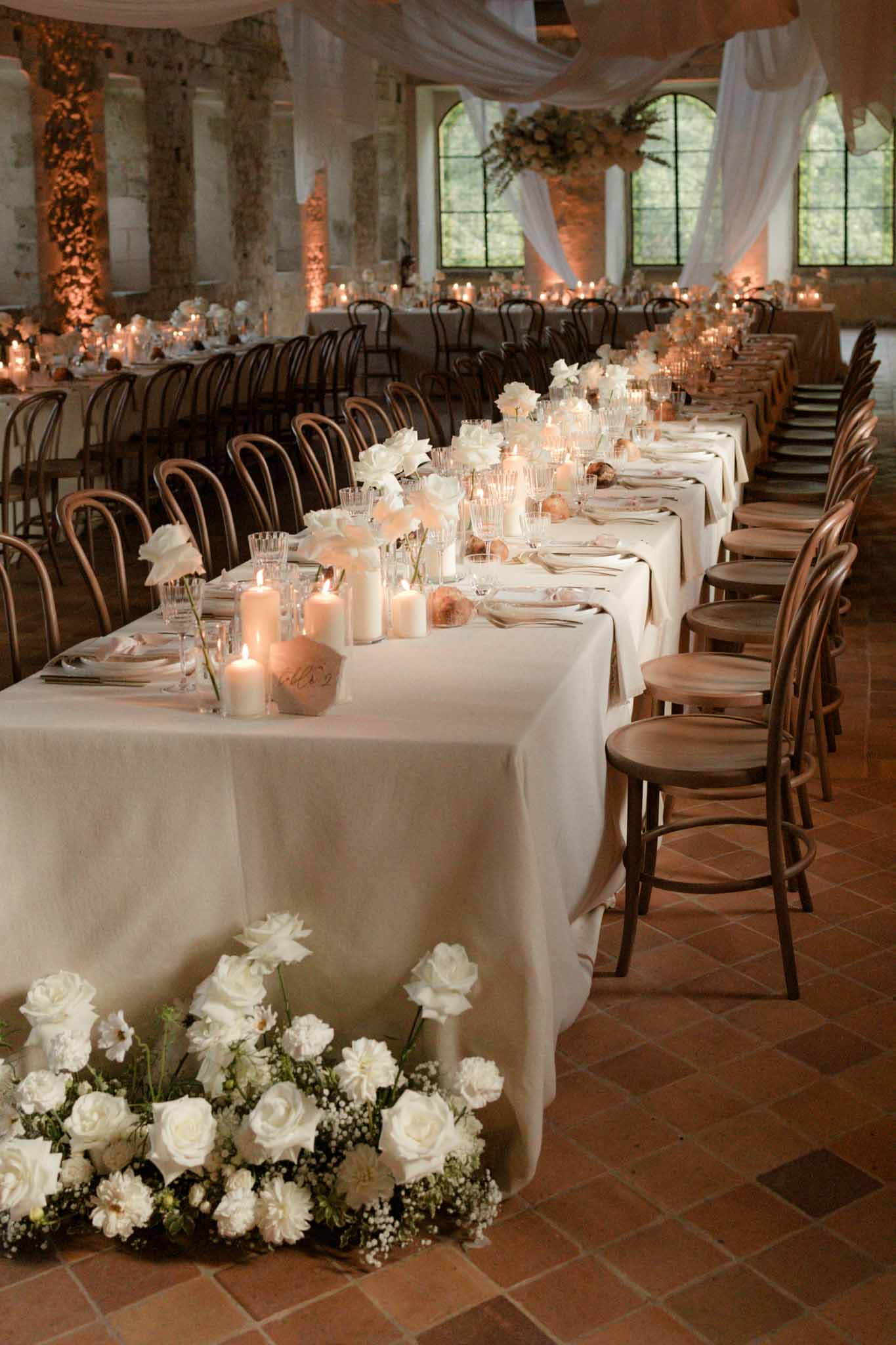 Long banquet tables with white roses and candles inside stone-walled venue with arched windows and wooden ceiling beams