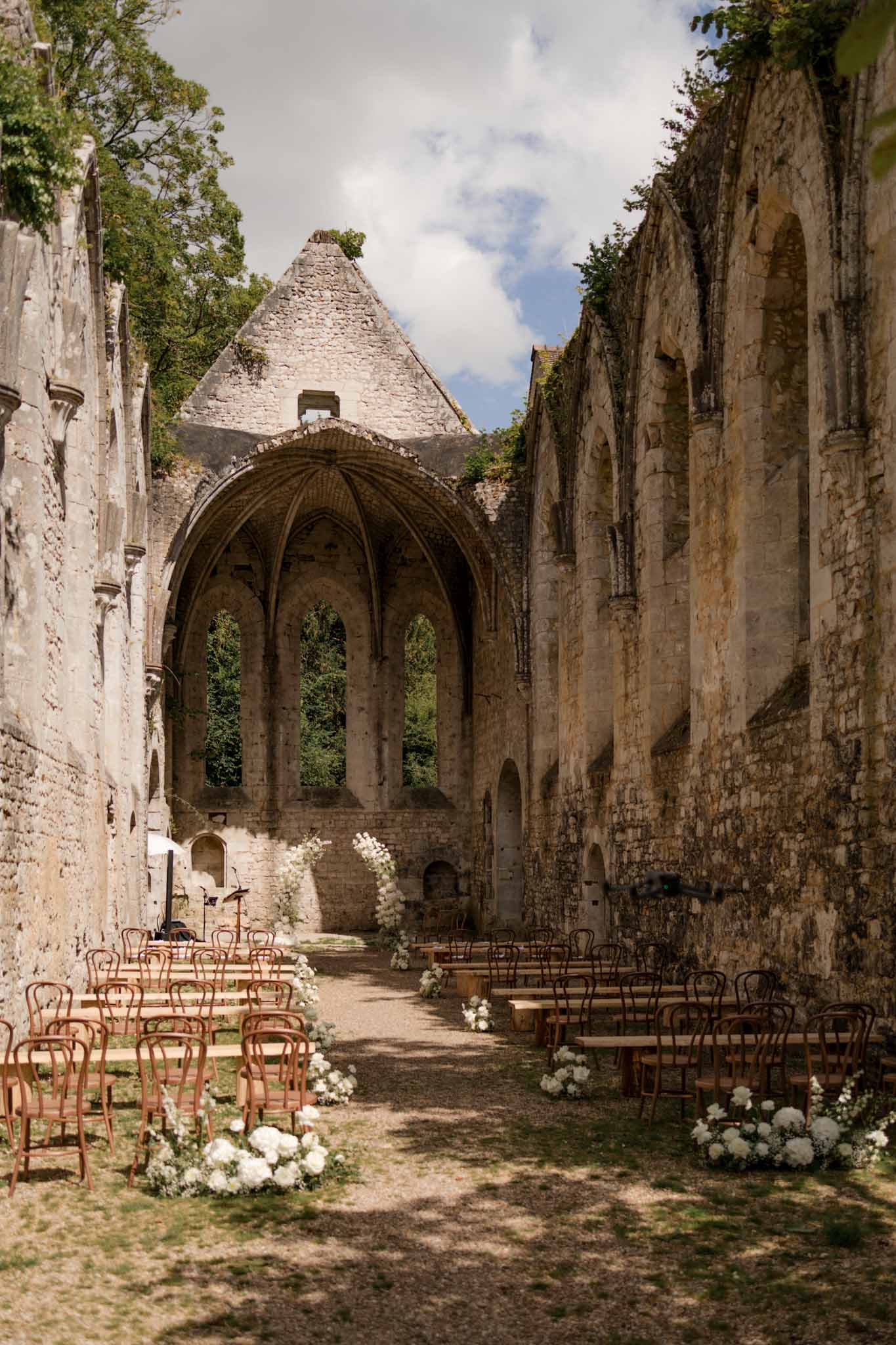 Wedding ceremony set up inside ruined medieval stone abbey with gothic arch, wooden chairs and white hydrangea aisle flowers