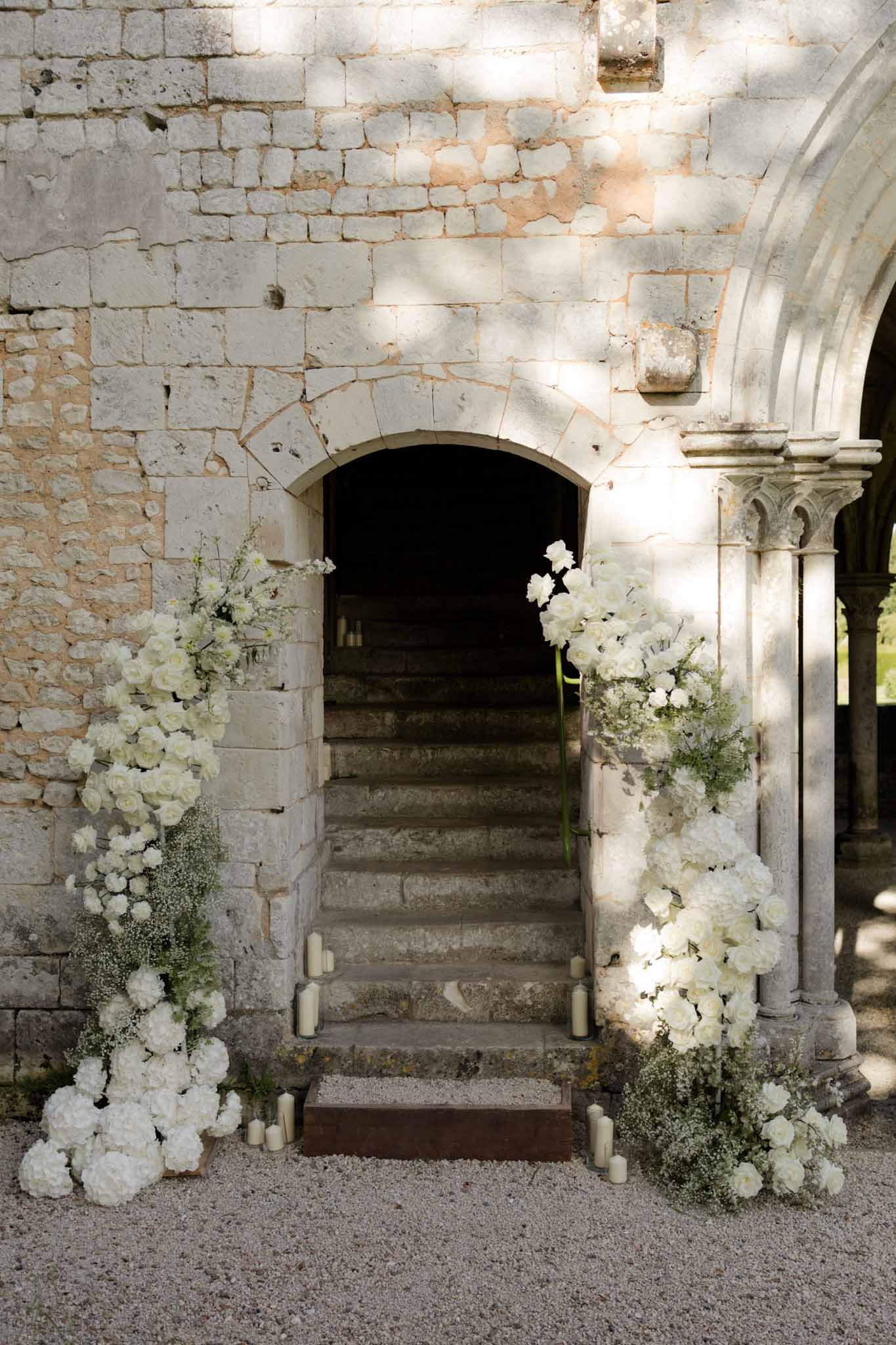 Stone archway with large white rose and hydrangea floral installations on pillars and ivory candles on steps