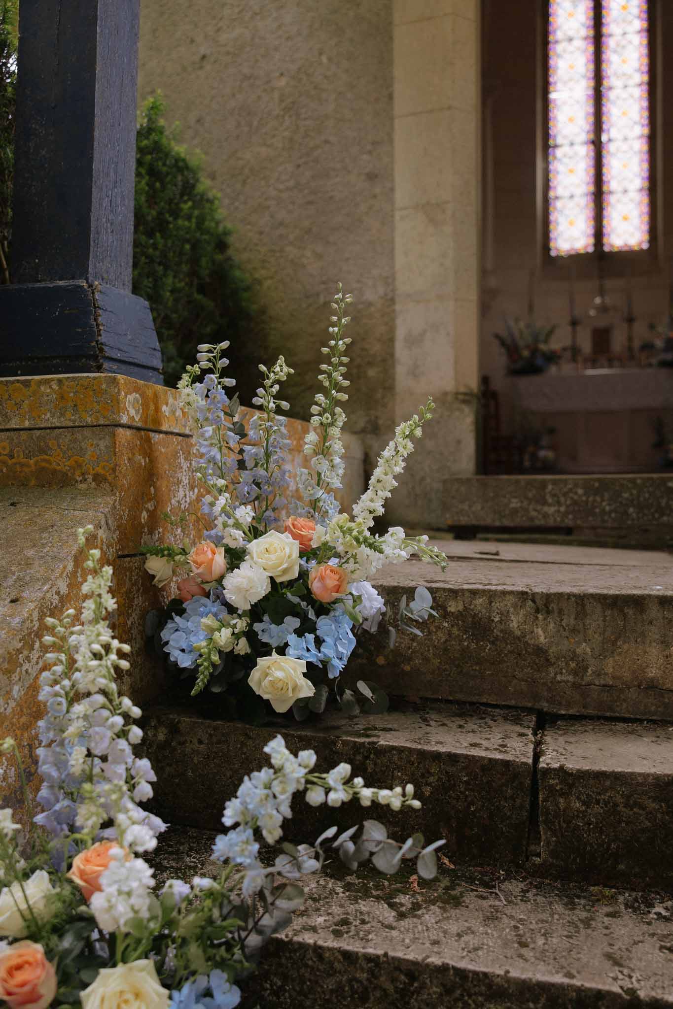Floral arrangements of ivory roses, peach roses, and blue hydrangeas on stone chapel steps with stained glass