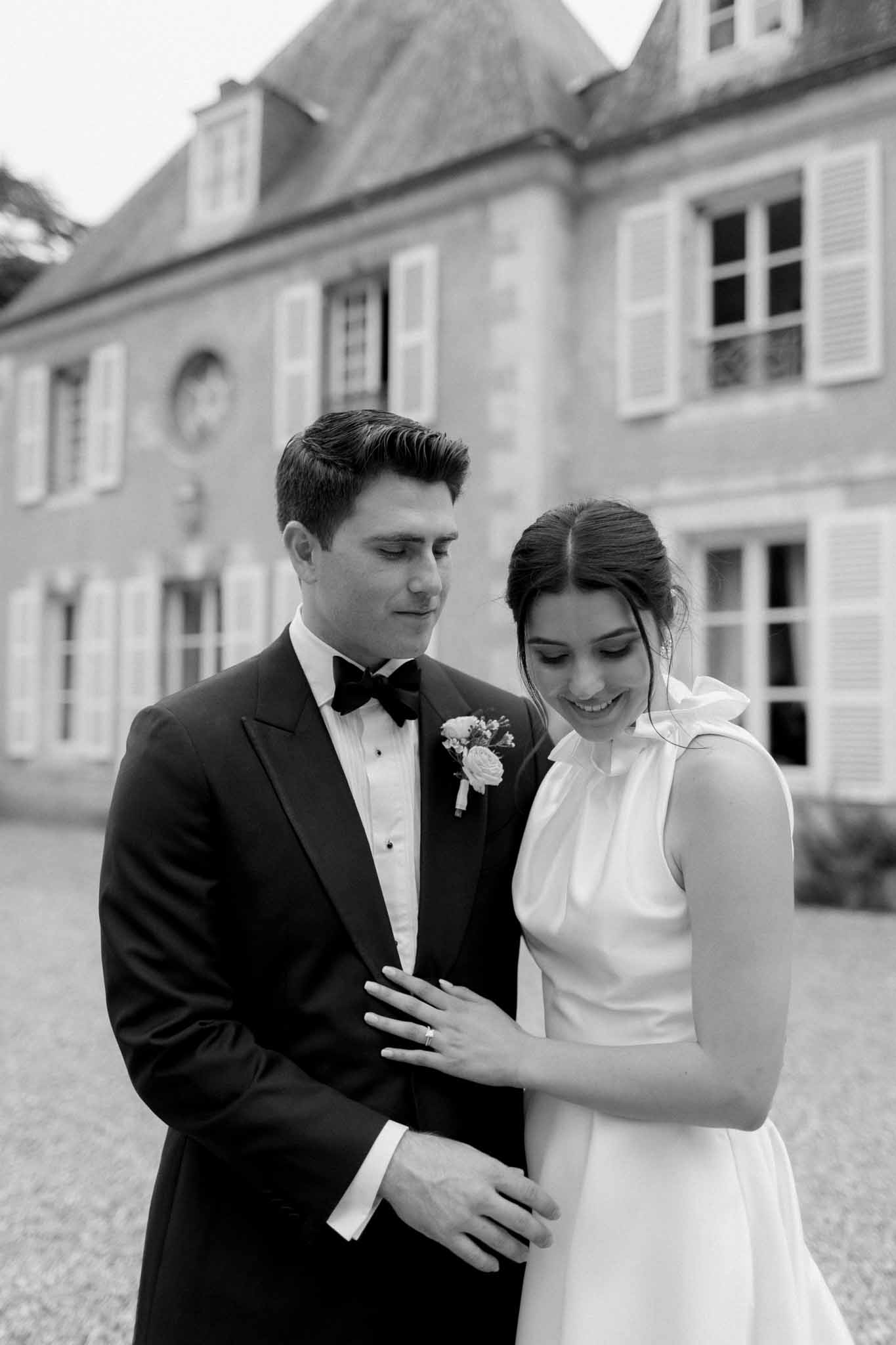 Black-and-white couple in courtyard before mansard-roofed manor, groom in tuxedo, bride in sleeveless white dress