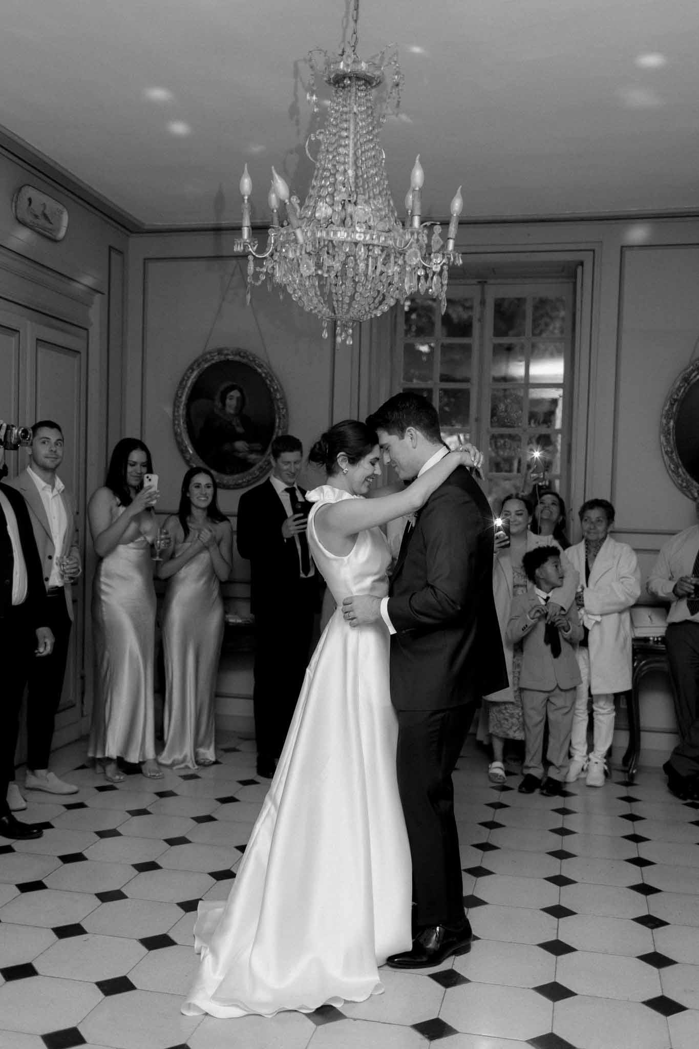 Black-and-white first dance on checkered ballroom floor beneath crystal chandelier with guests watching