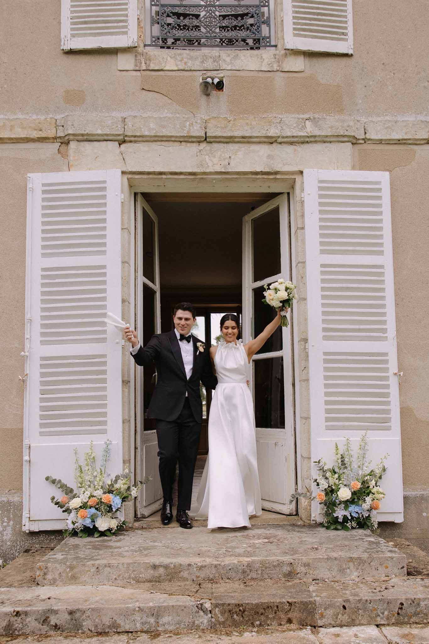 Bride and groom exiting stone building with bride holding bouquet overhead beside small floral arrangements