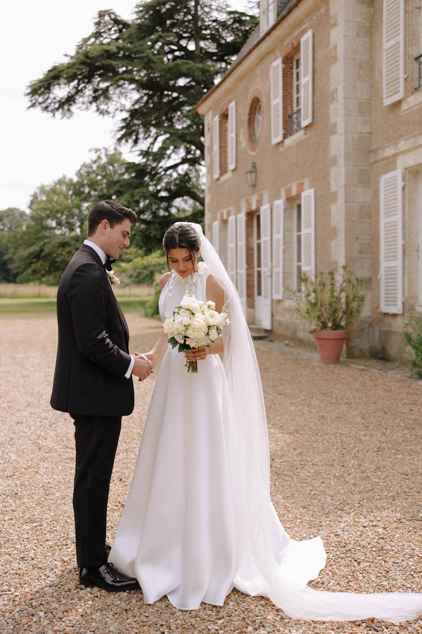 Bride and groom hold hands on gravel courtyard in front of stone manor house during first look
