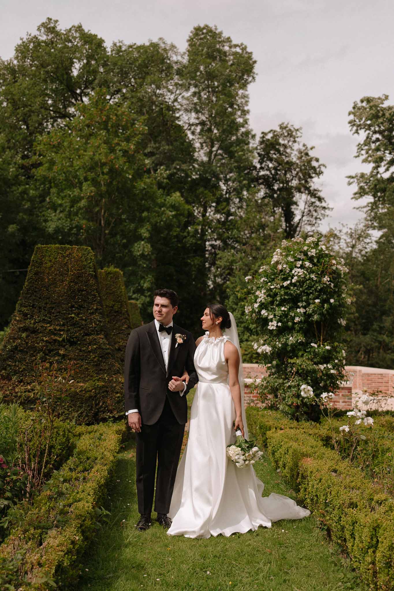 Bride and groom walking through formal garden with topiary hedges and white climbing rose archway