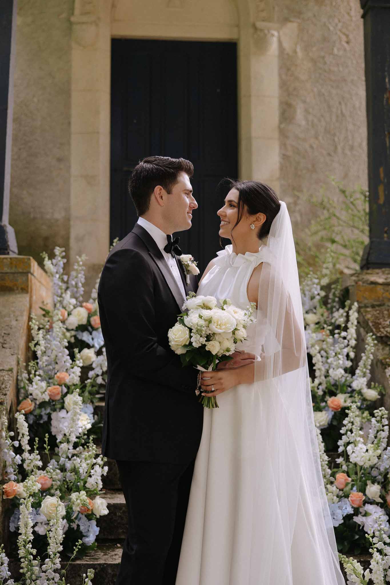 Bride in ivory silk gown and groom in black tuxedo facing each other outside stone chapel, flanked by white and peach florals