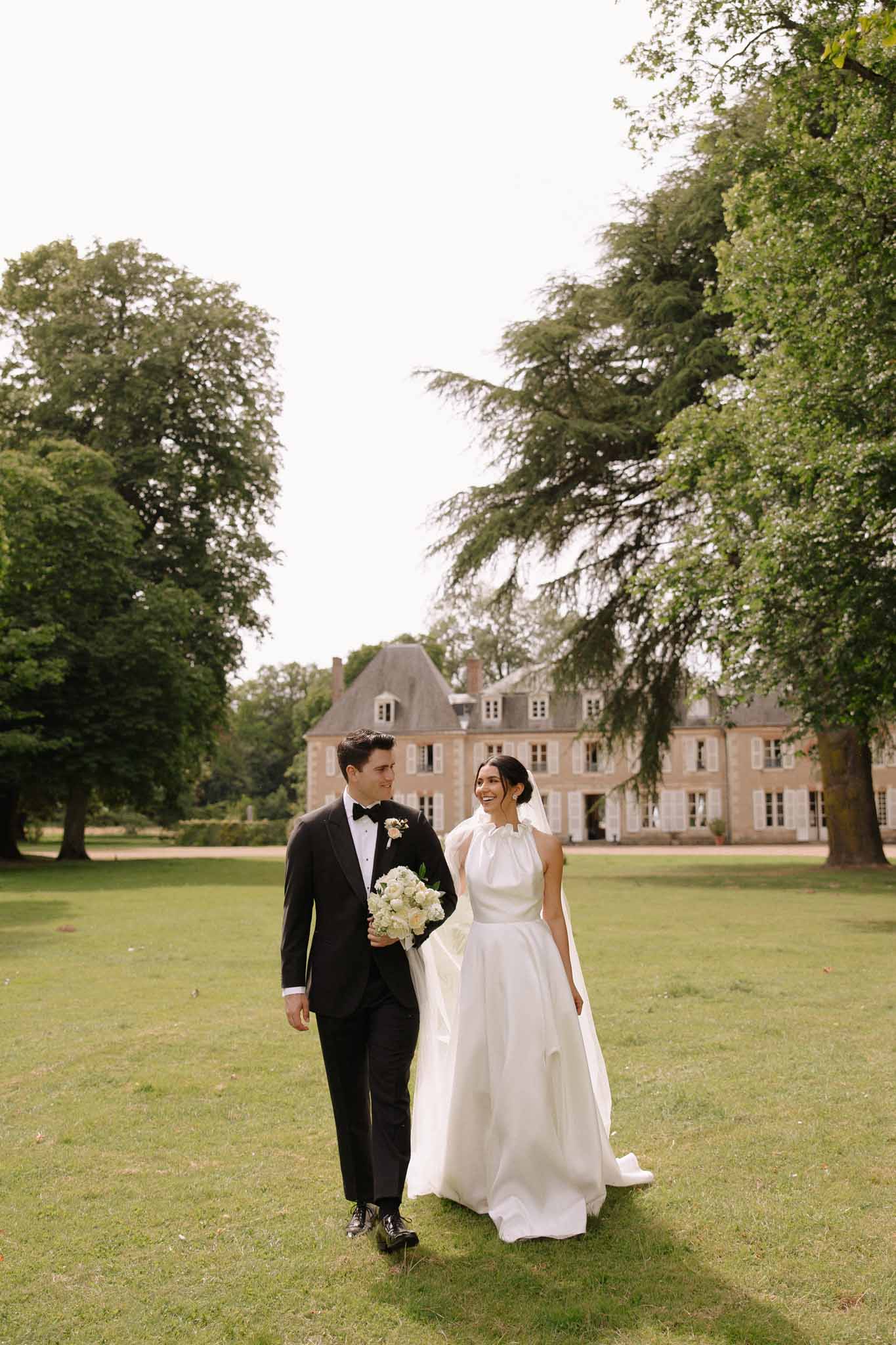 Bride in ivory gown and groom in tuxedo walking on manicured lawn in front of stone French chÃ¢teau