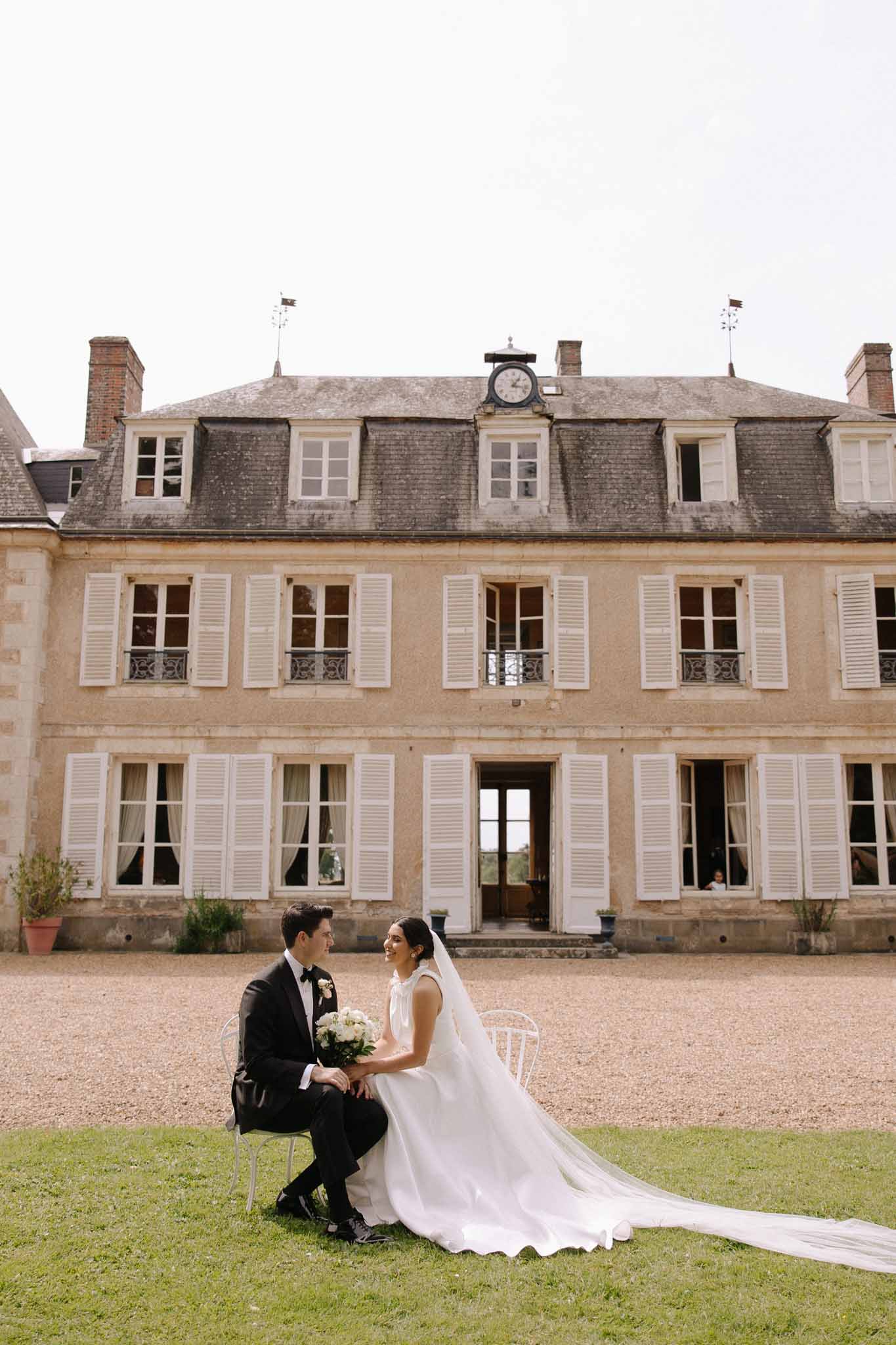 Bride and groom sit on white Chiavari chairs in front of three-story stone French manor with mansard roof and clock tower