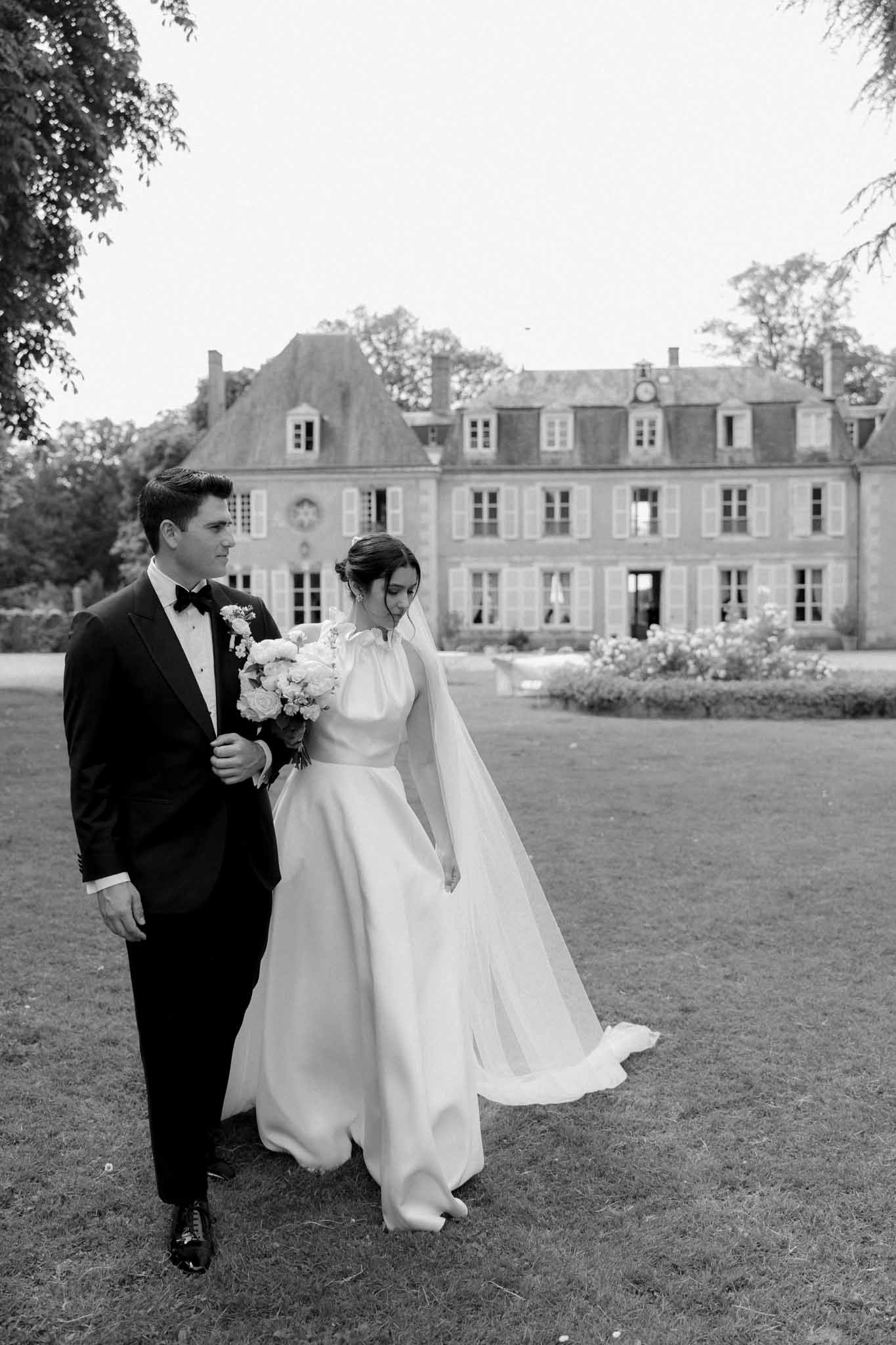 Black-and-white portrait of bride in long-sleeve gown with full skirt and groom on lawn before French chÃ¢teau