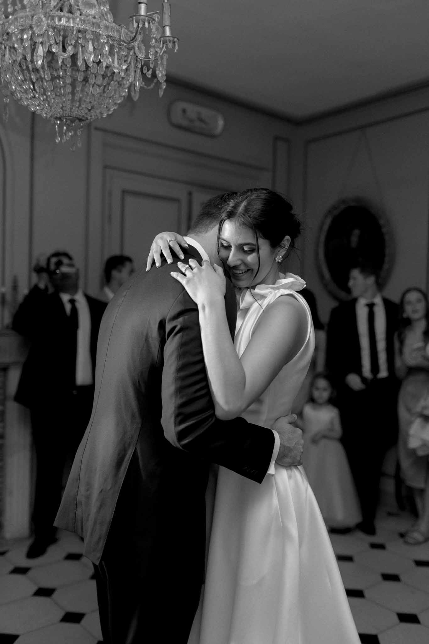 Black and white first dance in formal ballroom with crystal chandelier, guests watching from the edges