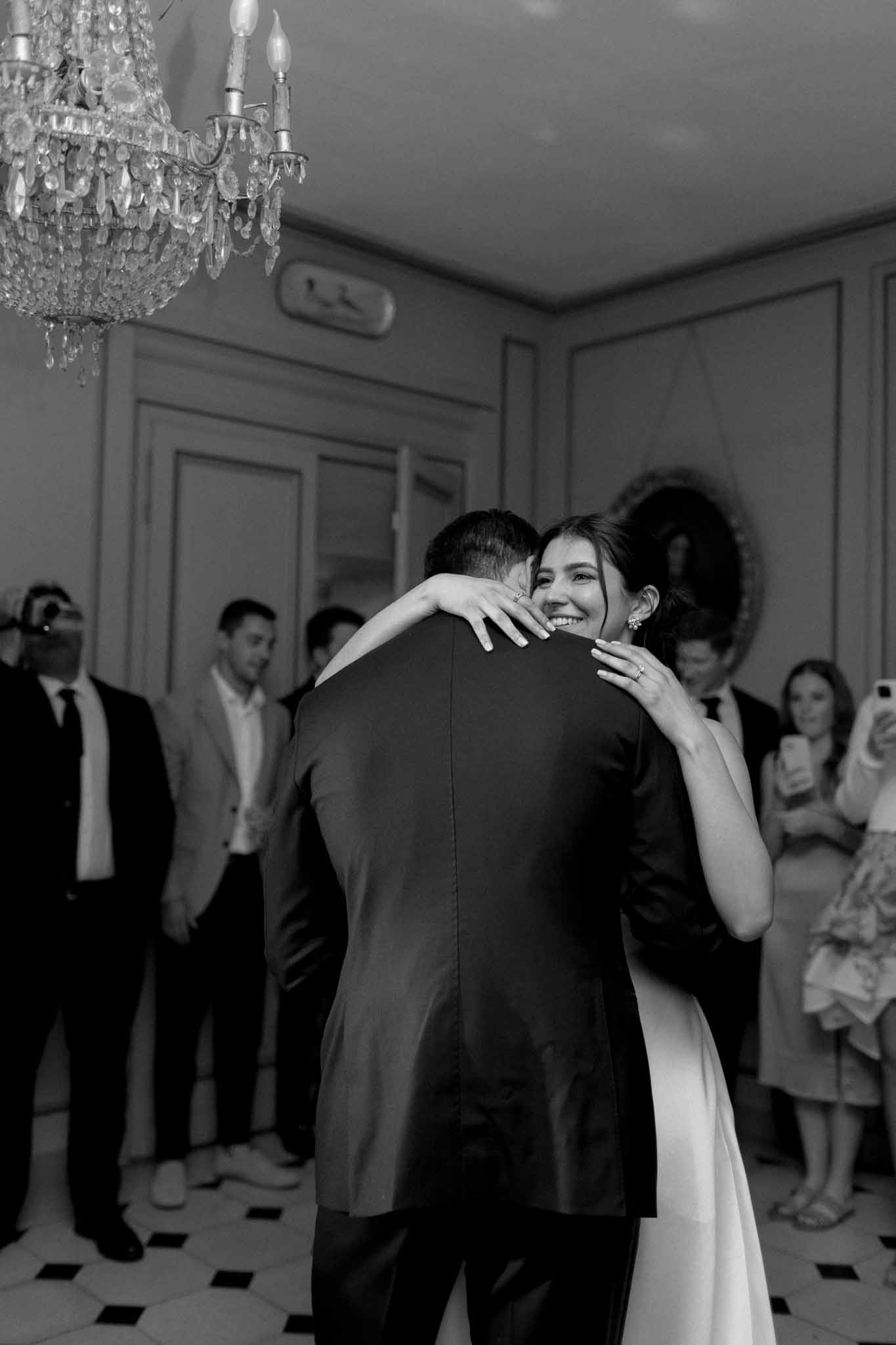 Black and white photo of couple's first dance under crystal chandelier in paneled ballroom