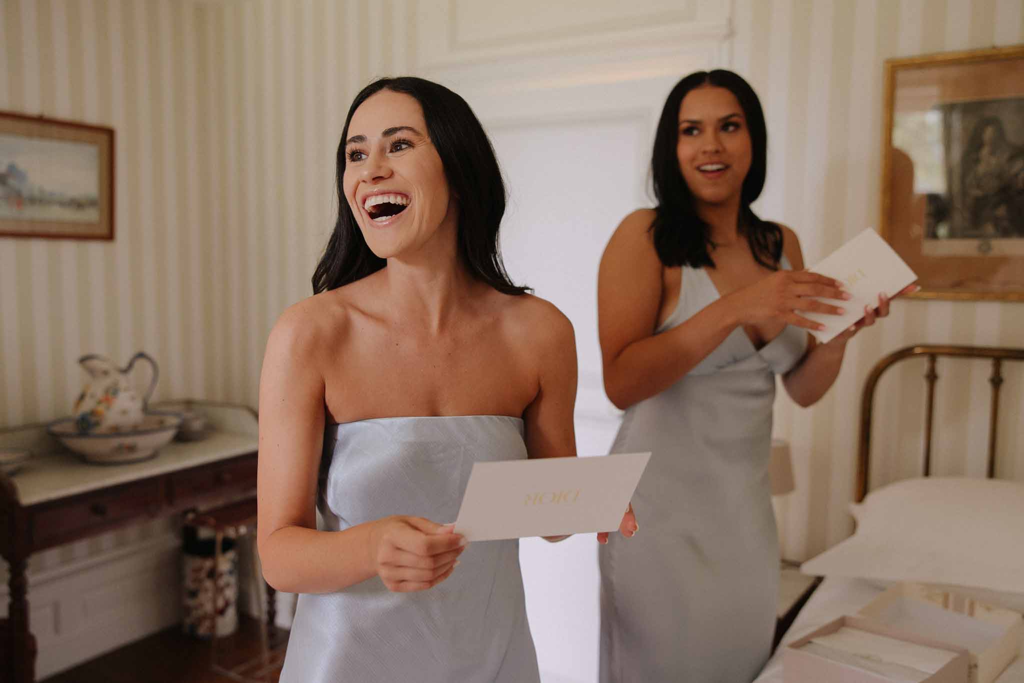 Two women in gray strapless dresses reading letters and laughing in an elegant interior room during wedding preparations.