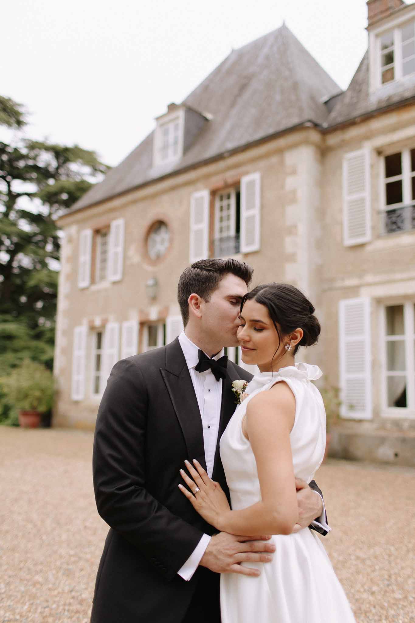 Bride and groom in courtyard of stone chÃ¢teau, groom in black tuxedo, bride in white A-line gown with bow detail